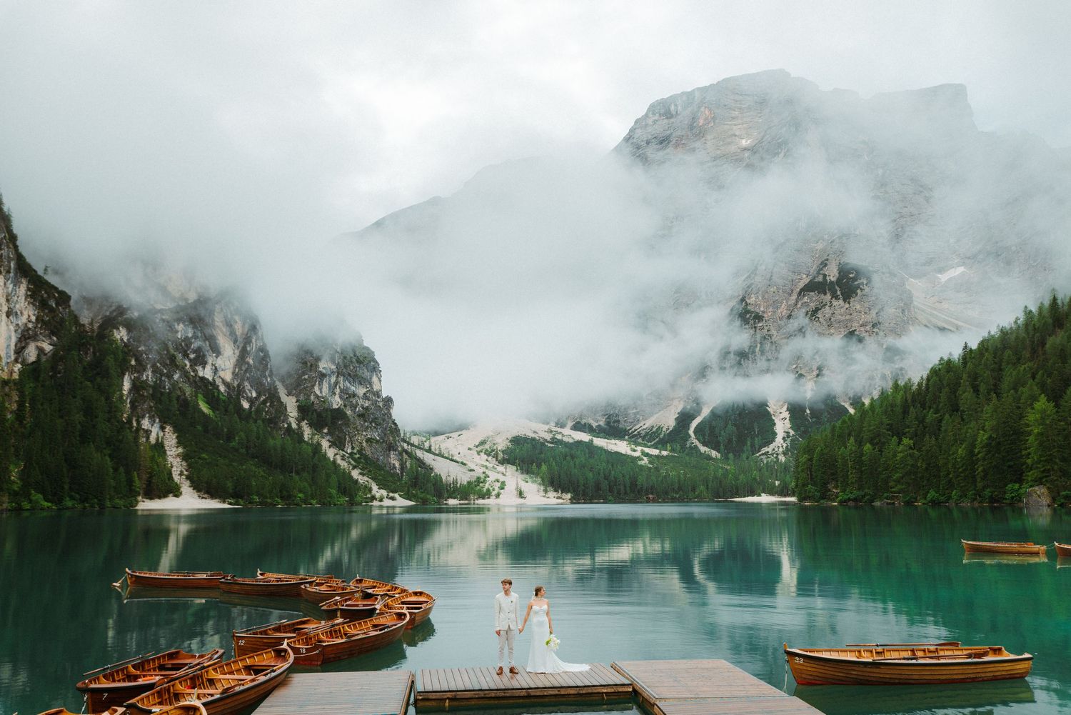 Wooden boats line the misty shore of Lake Braies in the Dolomites with fog-covered mountains in the background.
