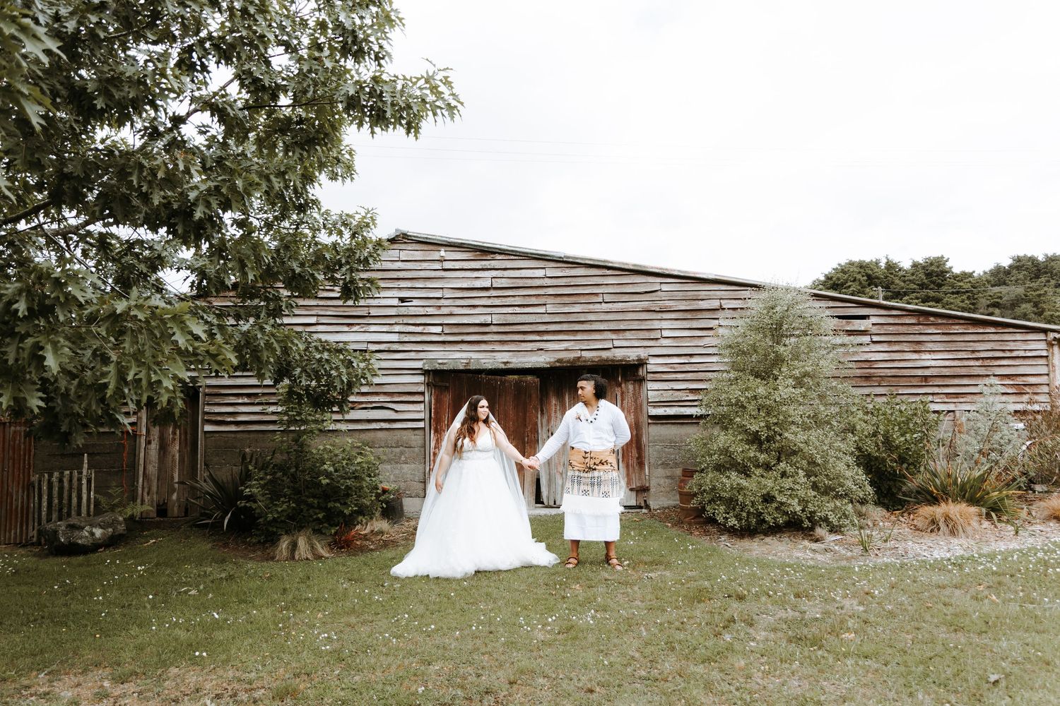 A rustic outdoor wedding scene with people in white attire standing in front of a wooden barn structure.