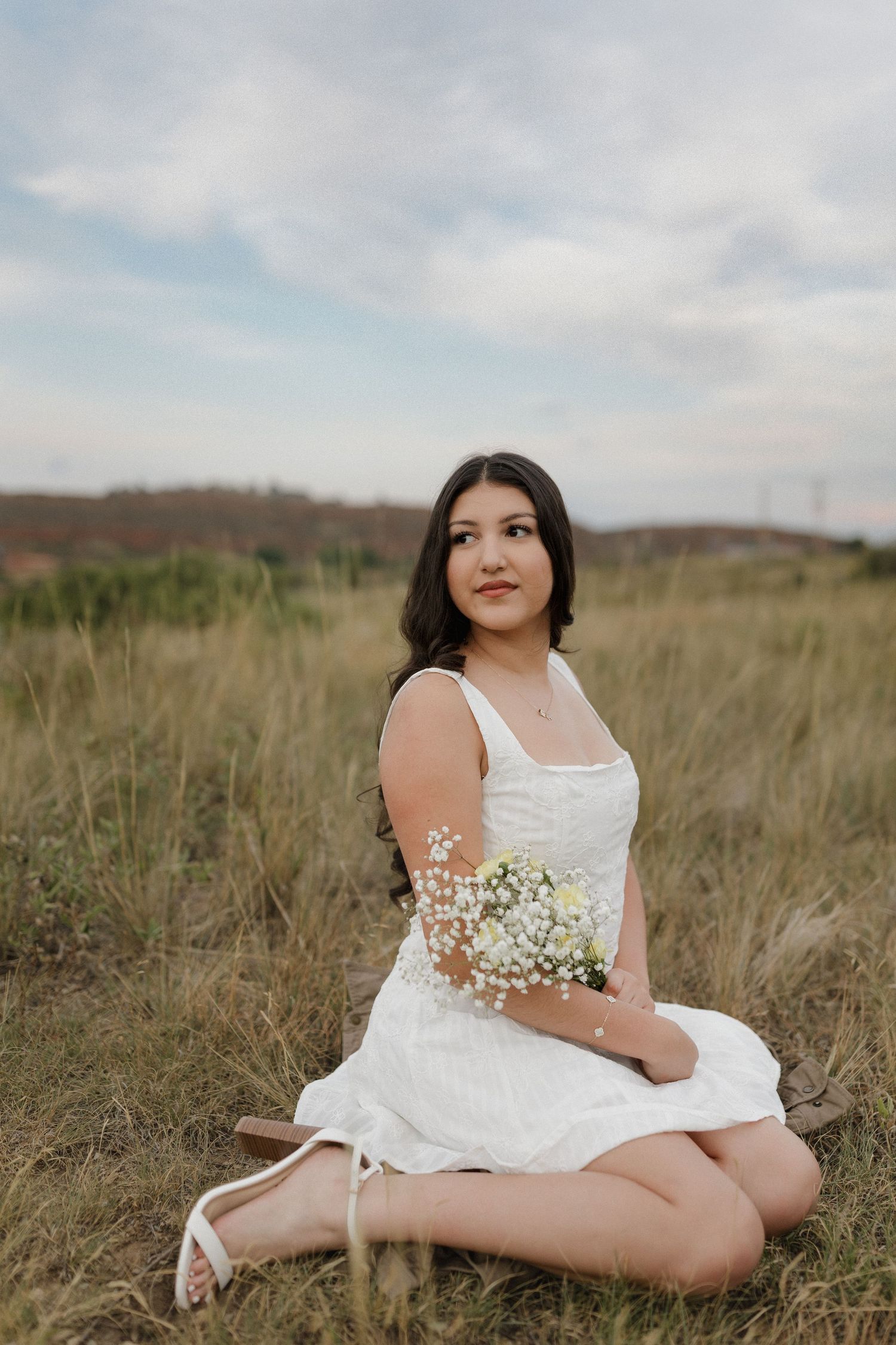 A white dress-clad figure reclines in a serene field of tall grass while holding delicate baby's breath flowers.