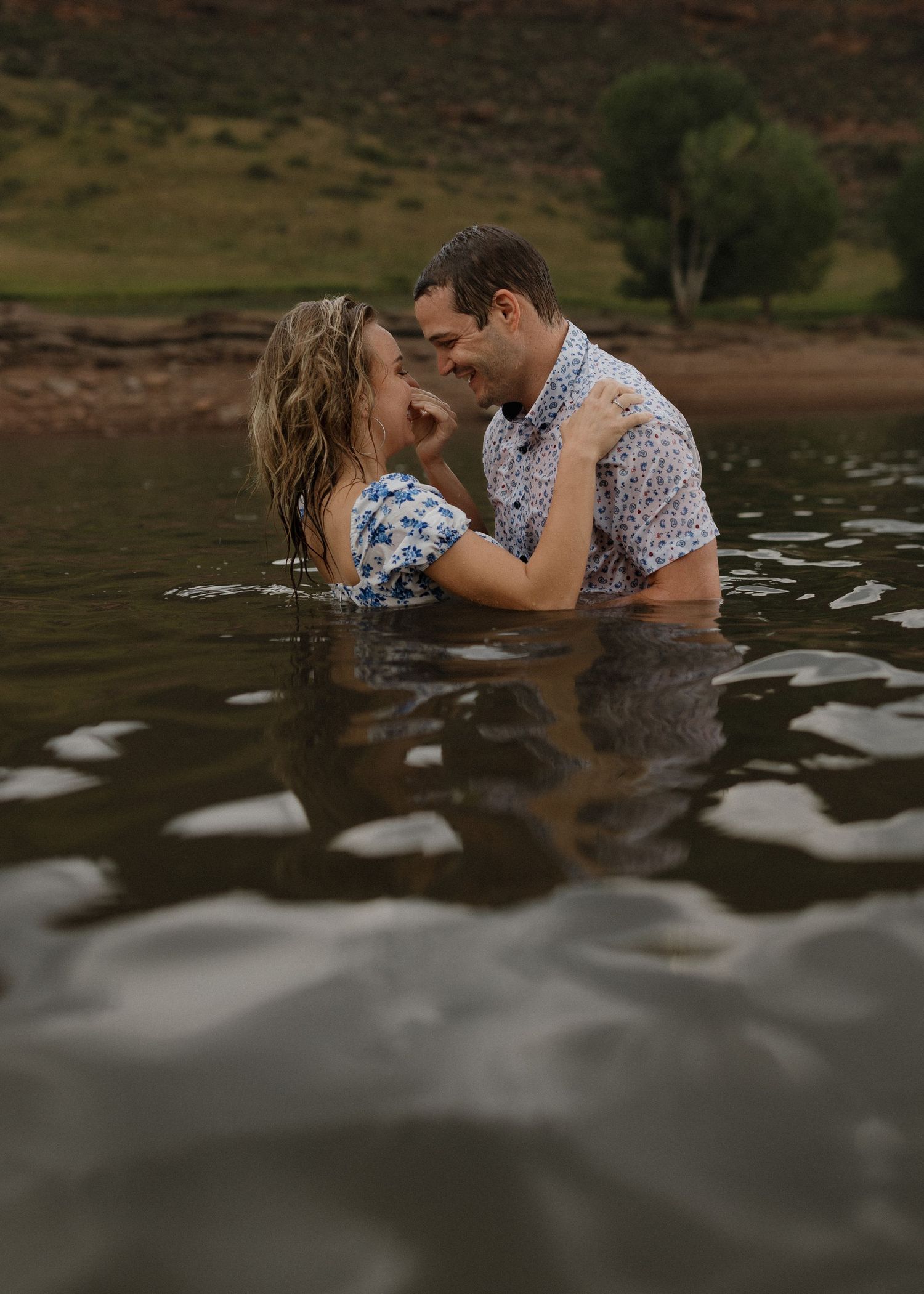 A couple embraces while standing in shallow water against a scenic natural backdrop at sunset.