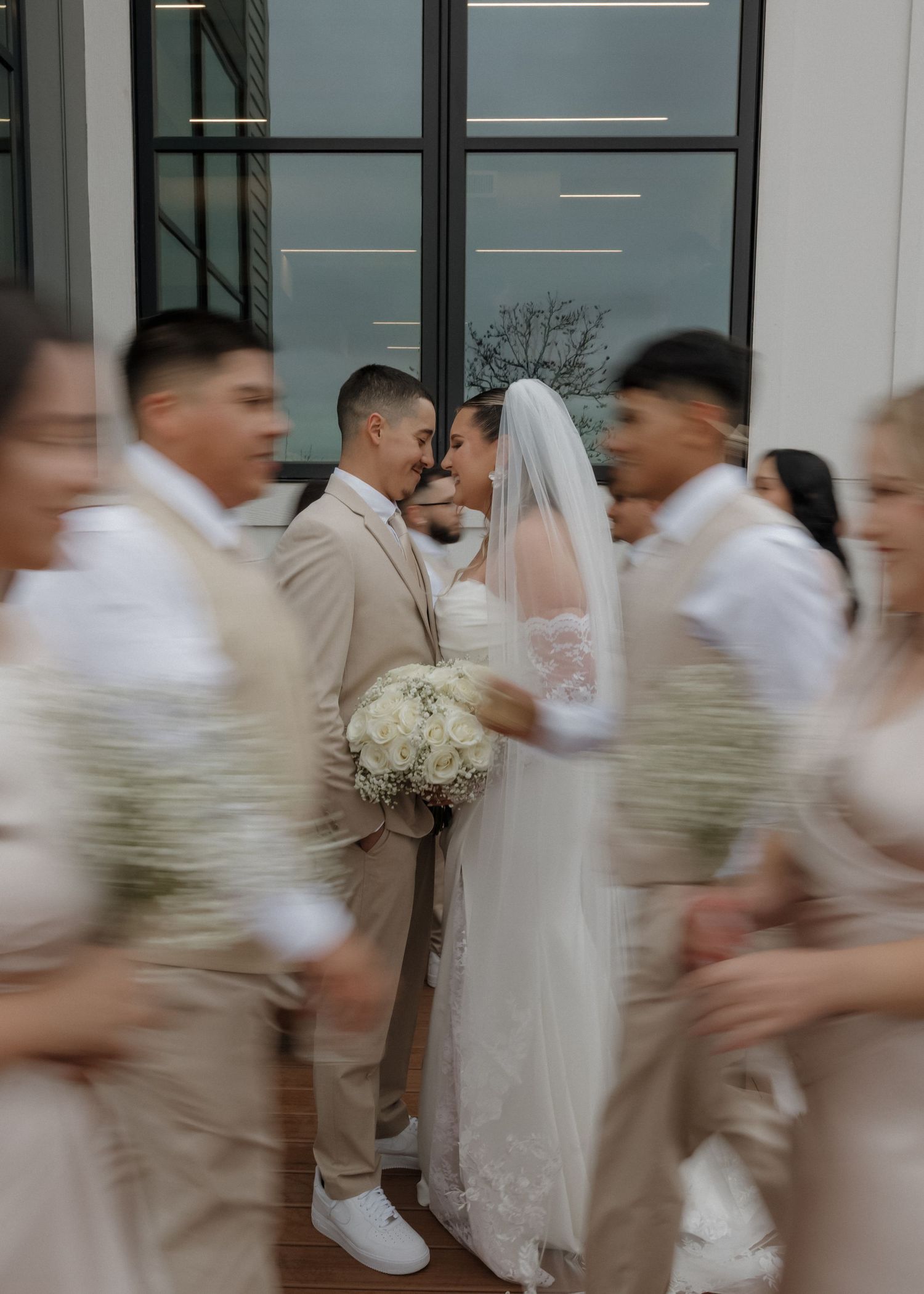 Wedding party in motion blur with groomsmen in beige suits and bridesmaids in white dresses moving down a hallway.