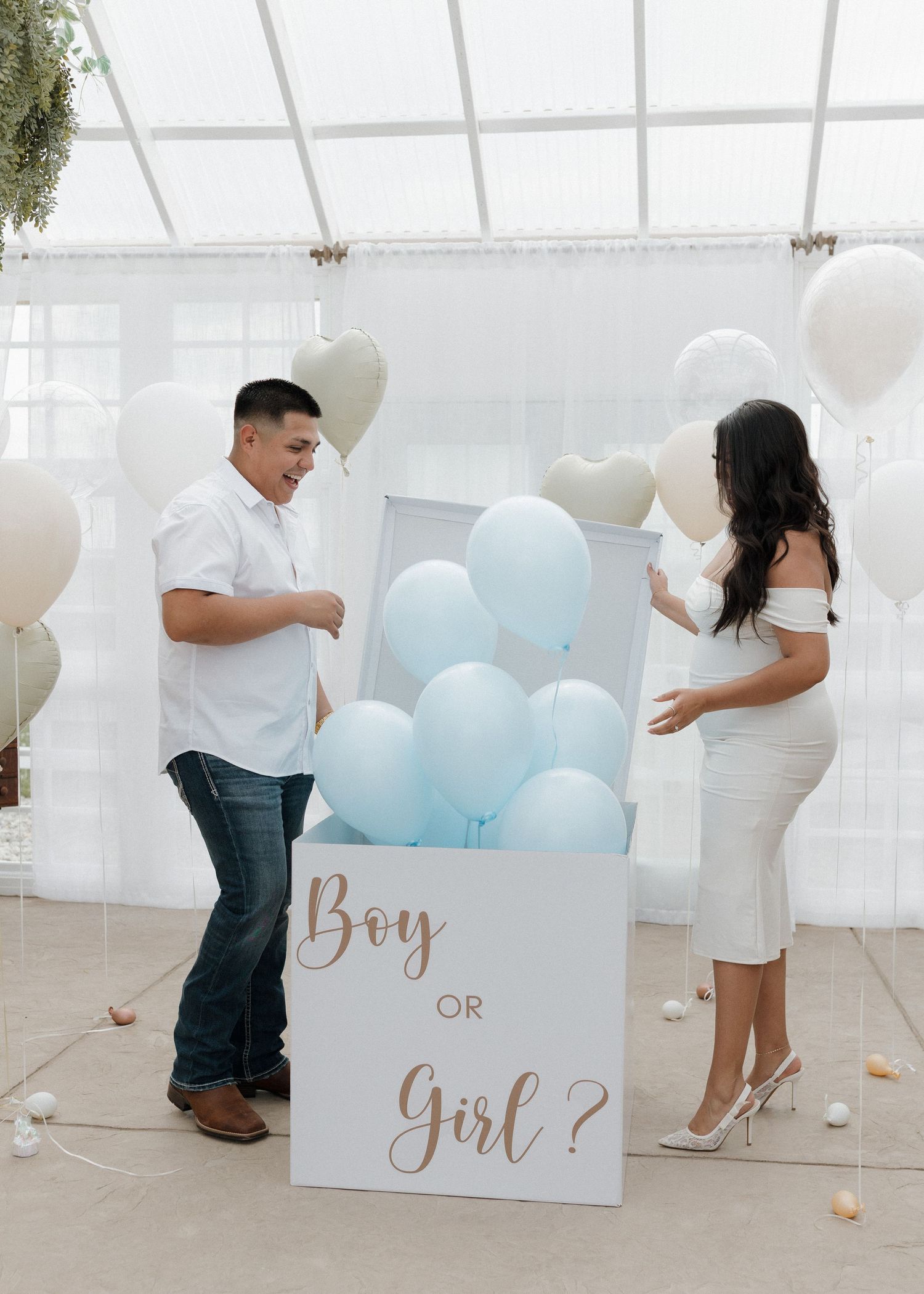 Two people stand next to a box with blue and white balloons at a gender reveal party setup in a bright indoor space.