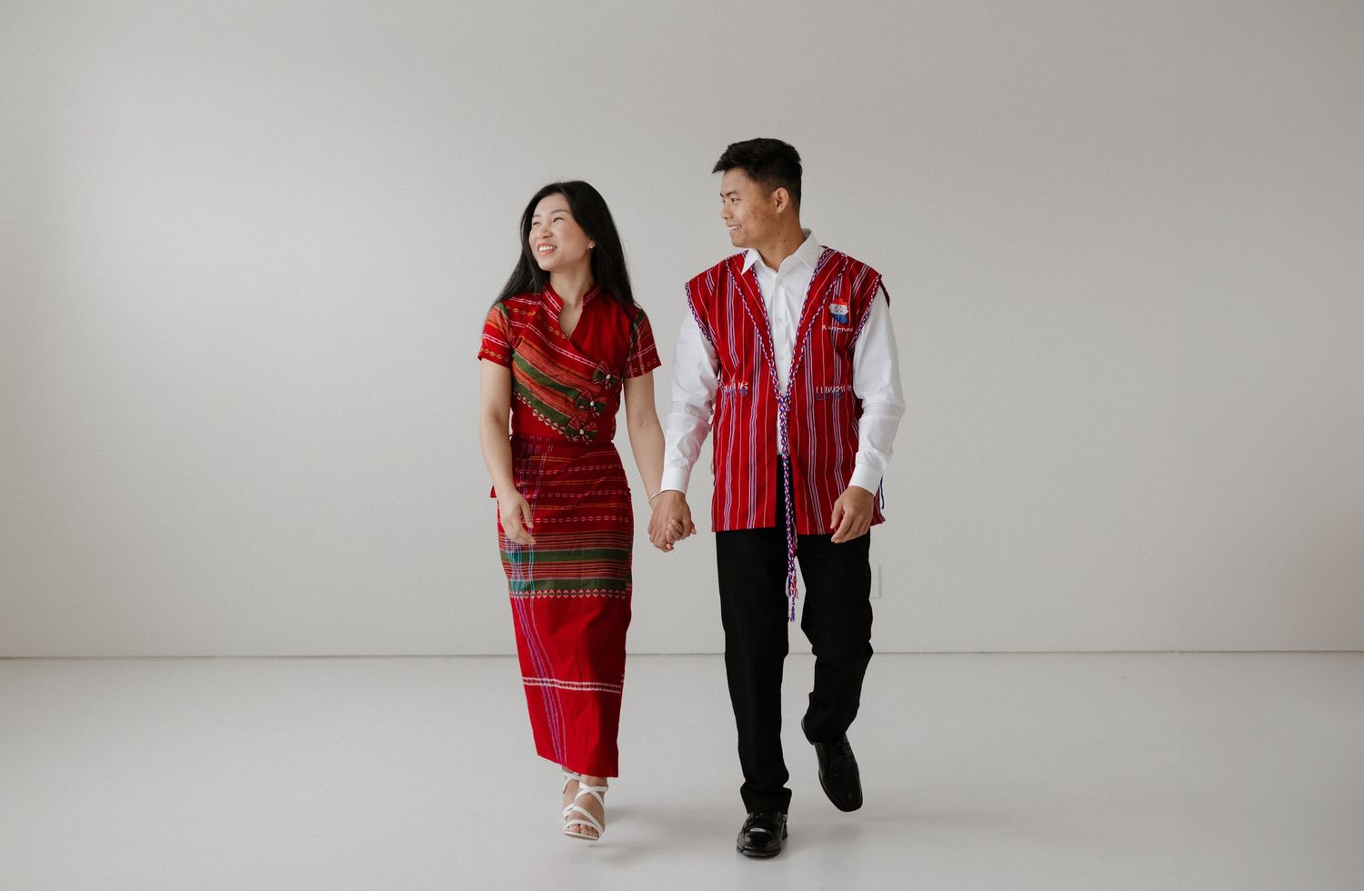 Two people wearing traditional red Asian-inspired clothing walk hand in hand against a white studio backdrop.