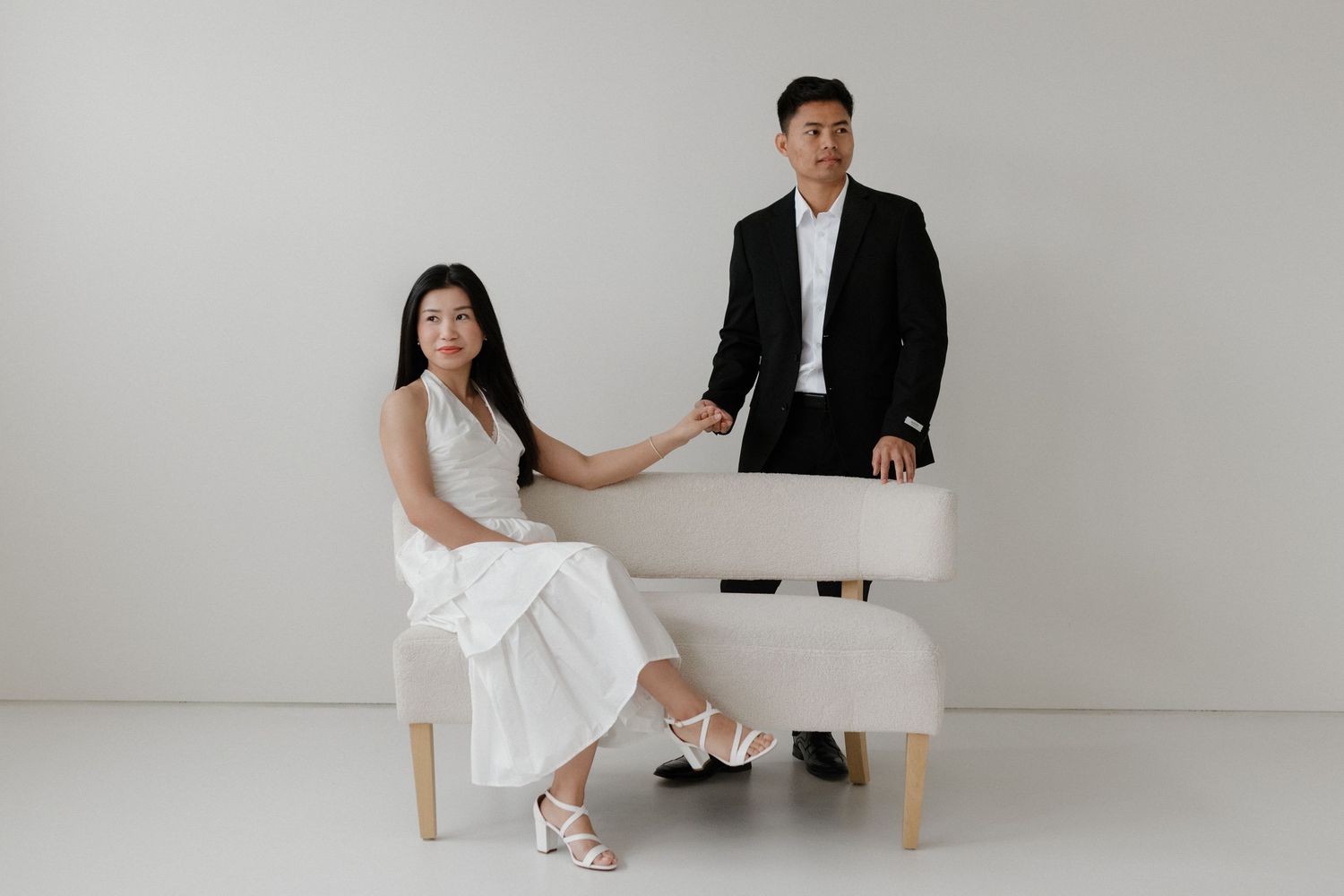 Couple in formal attire poses with white piano against minimalist white backdrop for elegant studio photoshoot.