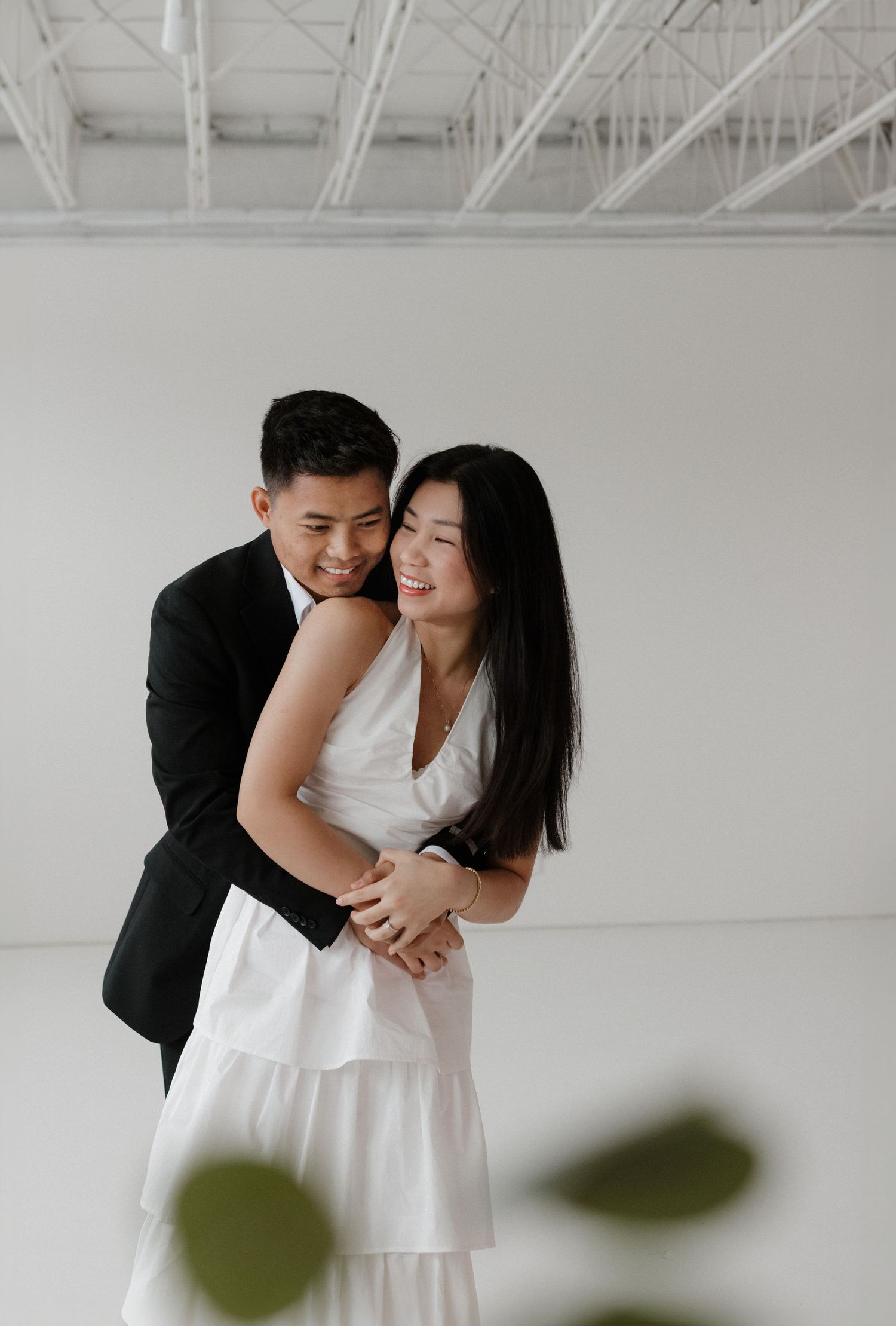 A joyful couple embraces and laughs together in a bright white studio setting with industrial ceiling.
