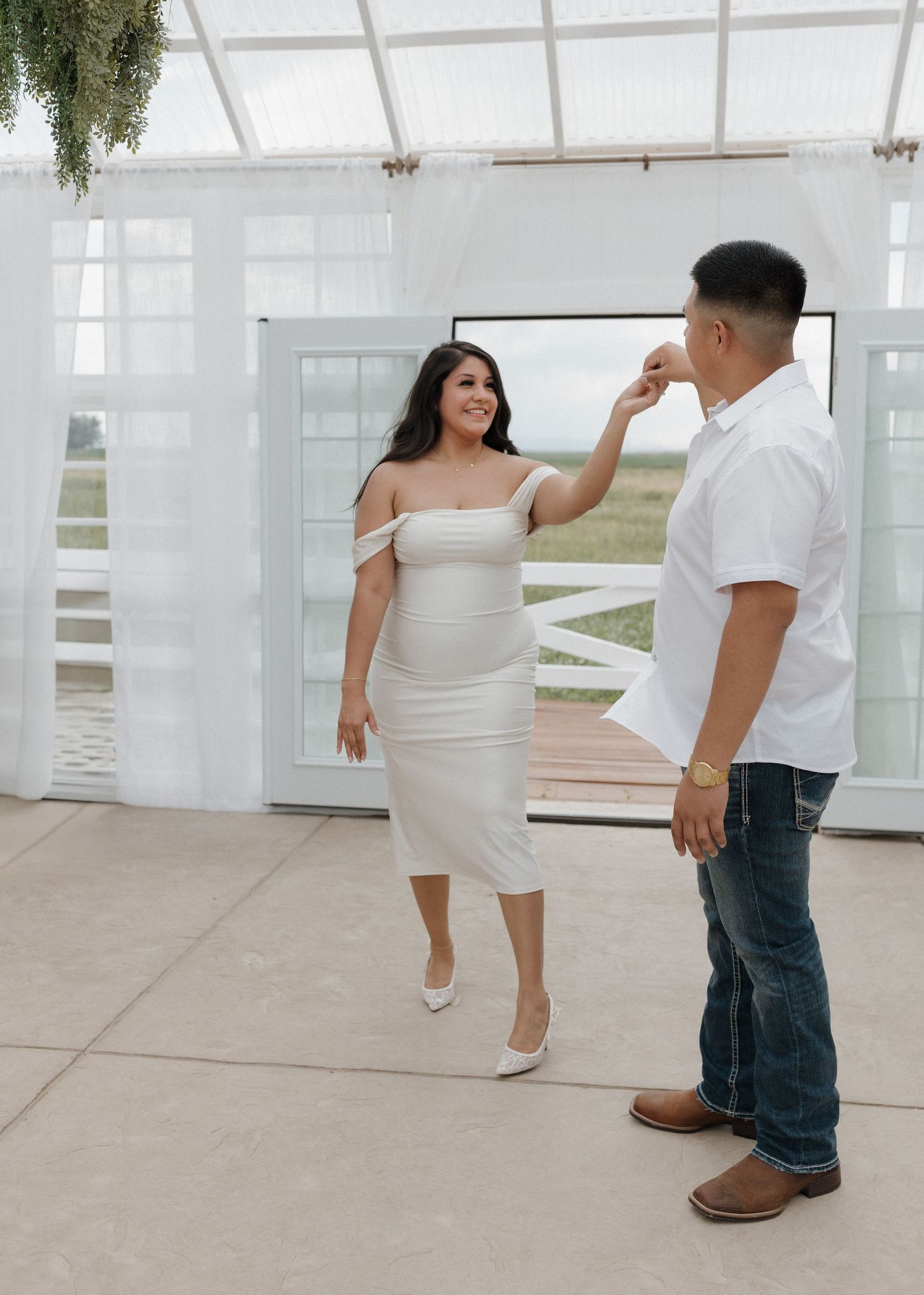Two people share a romantic moment in a modern white venue with glass doors and white sheer curtains.