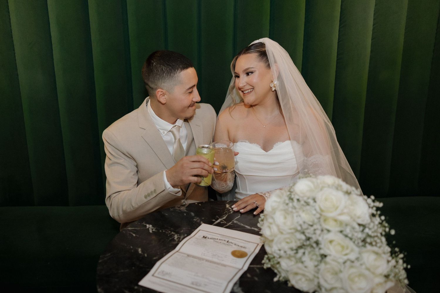 A newlywed couple sharing a drink while sitting in front of a green velvet backdrop with a white floral bouquet nearby.