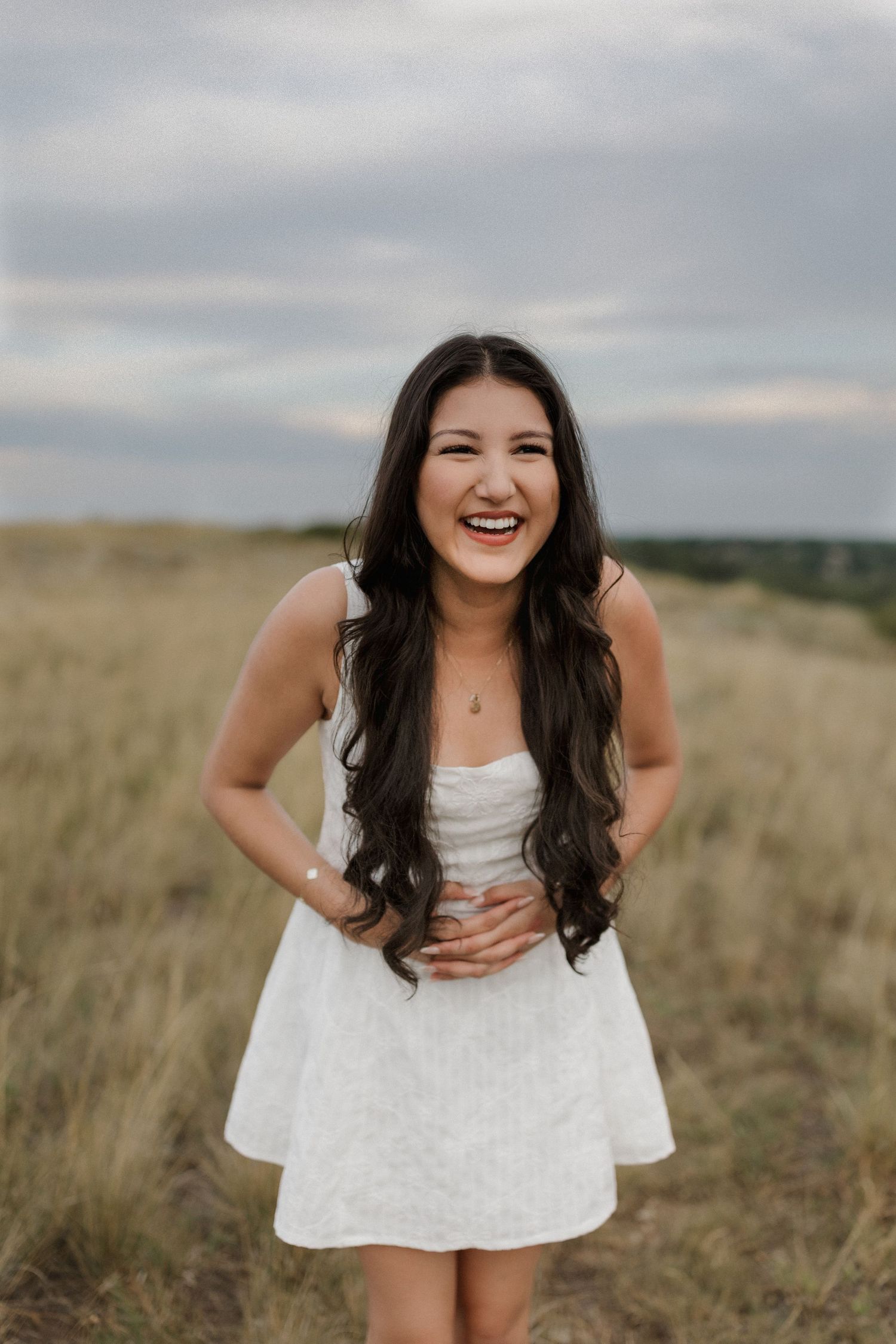 Person in white dress smiling and posing in a golden grass field on an overcast day.