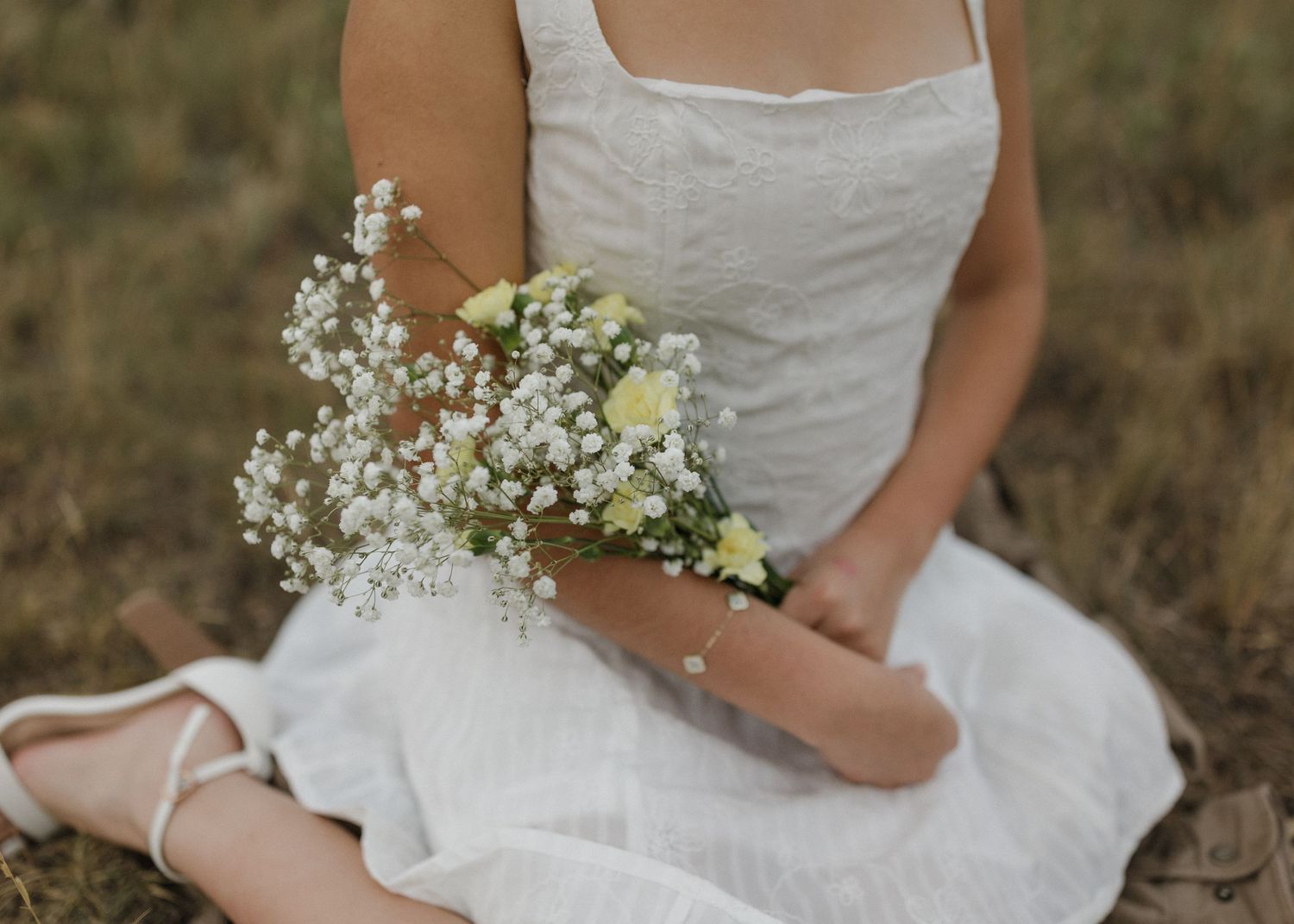 Someone in a white dress sits outdoors holding a delicate bouquet of baby's breath and yellow flowers.