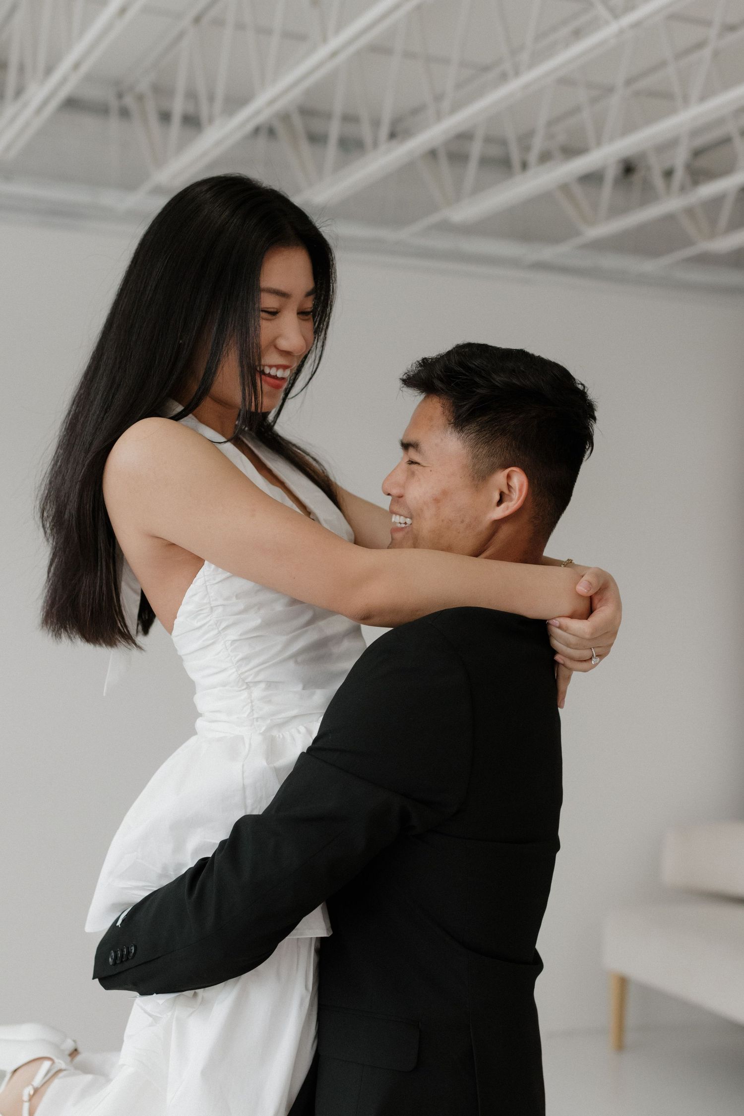Young couple embracing in a white studio space, with her in a white dress and him in a black suit.