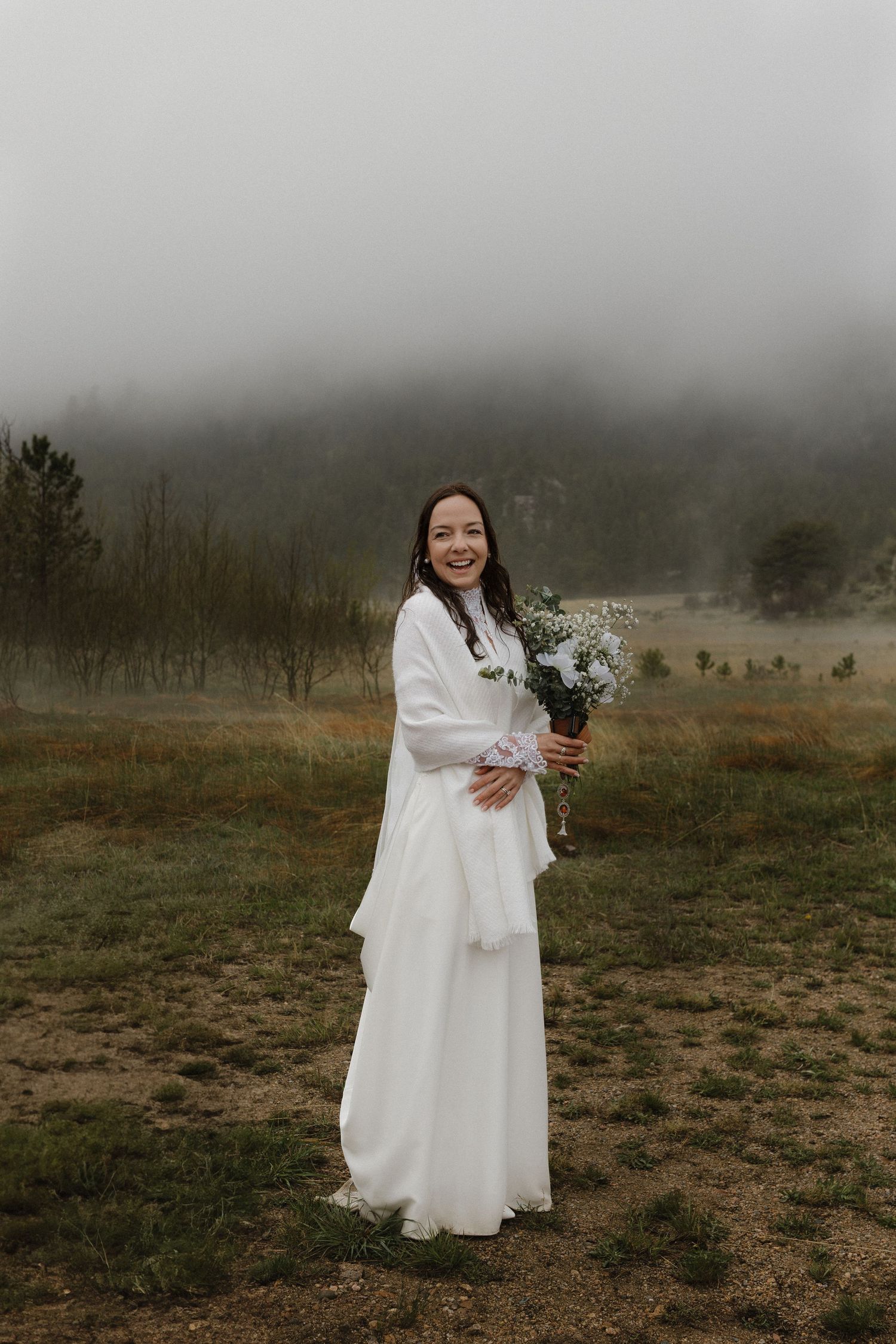 A person in a white wedding dress and wrap holds a bouquet while standing in a misty mountain landscape.