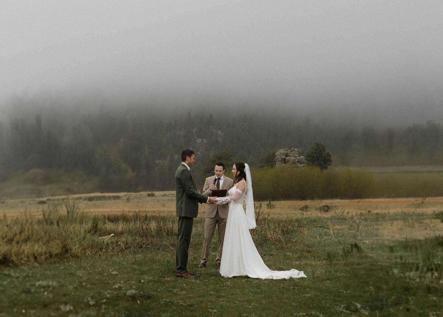 A couple stands in a misty field wearing wedding attire during a foggy outdoor ceremony.