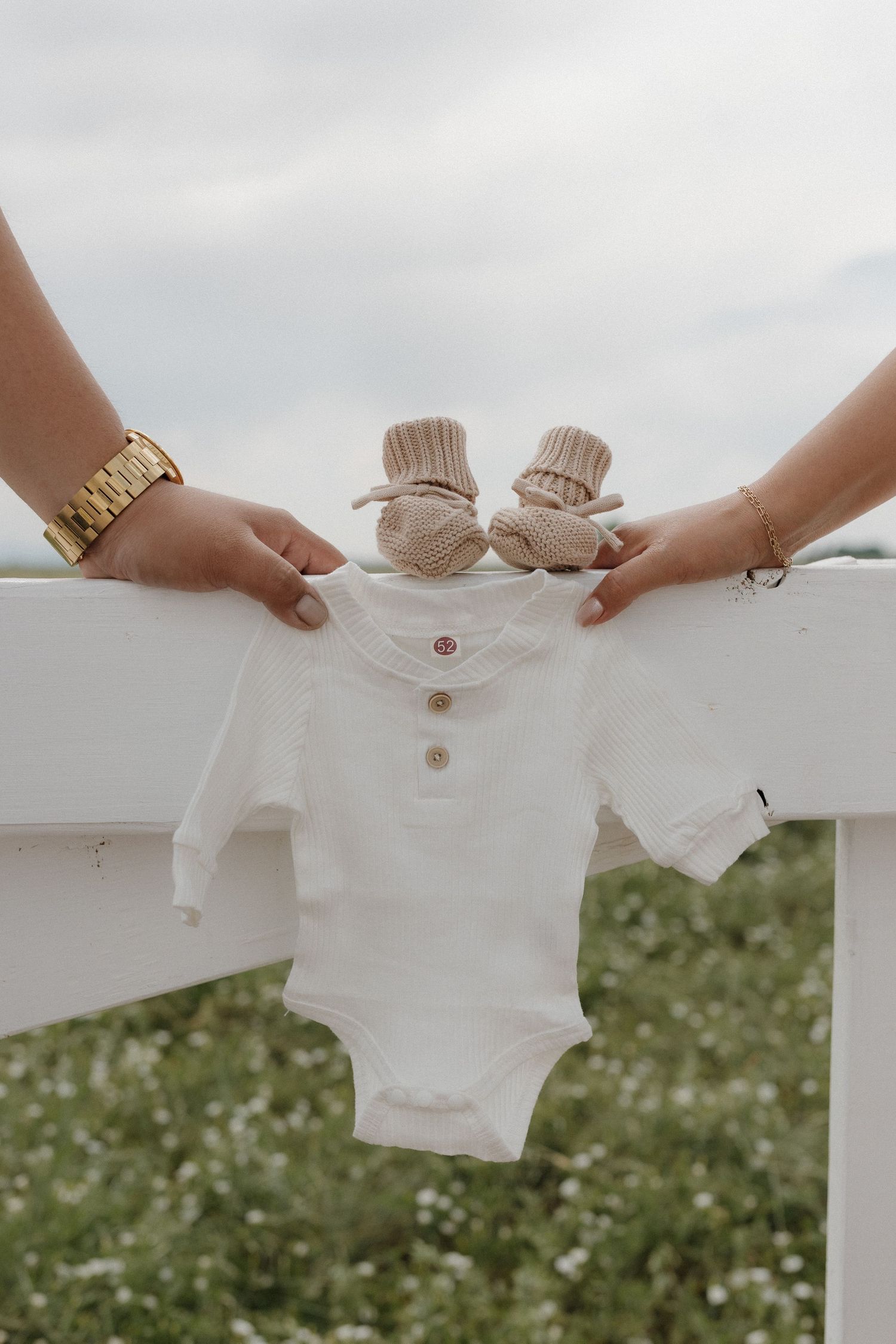 Four identical white baby onesies lined up in a row against a blurred grassy background with white flowers.