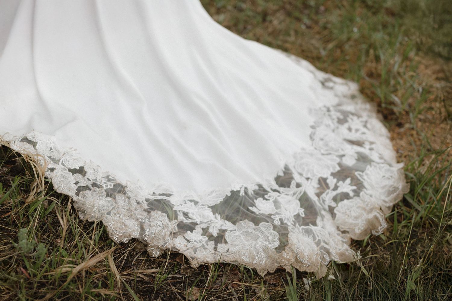 A white wedding dress train with delicate lace details trails gracefully across green grass in an outdoor setting.