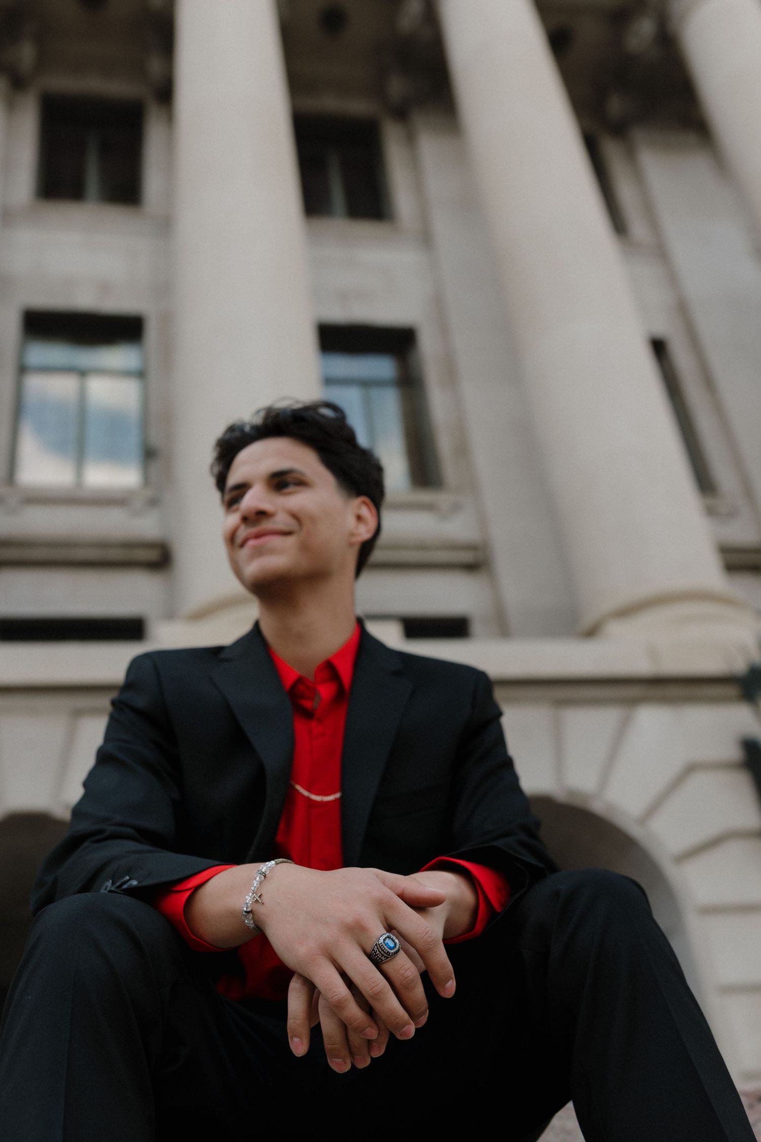 Portrait of someone in a black suit with red tie sitting on steps with white classical columns in the background.