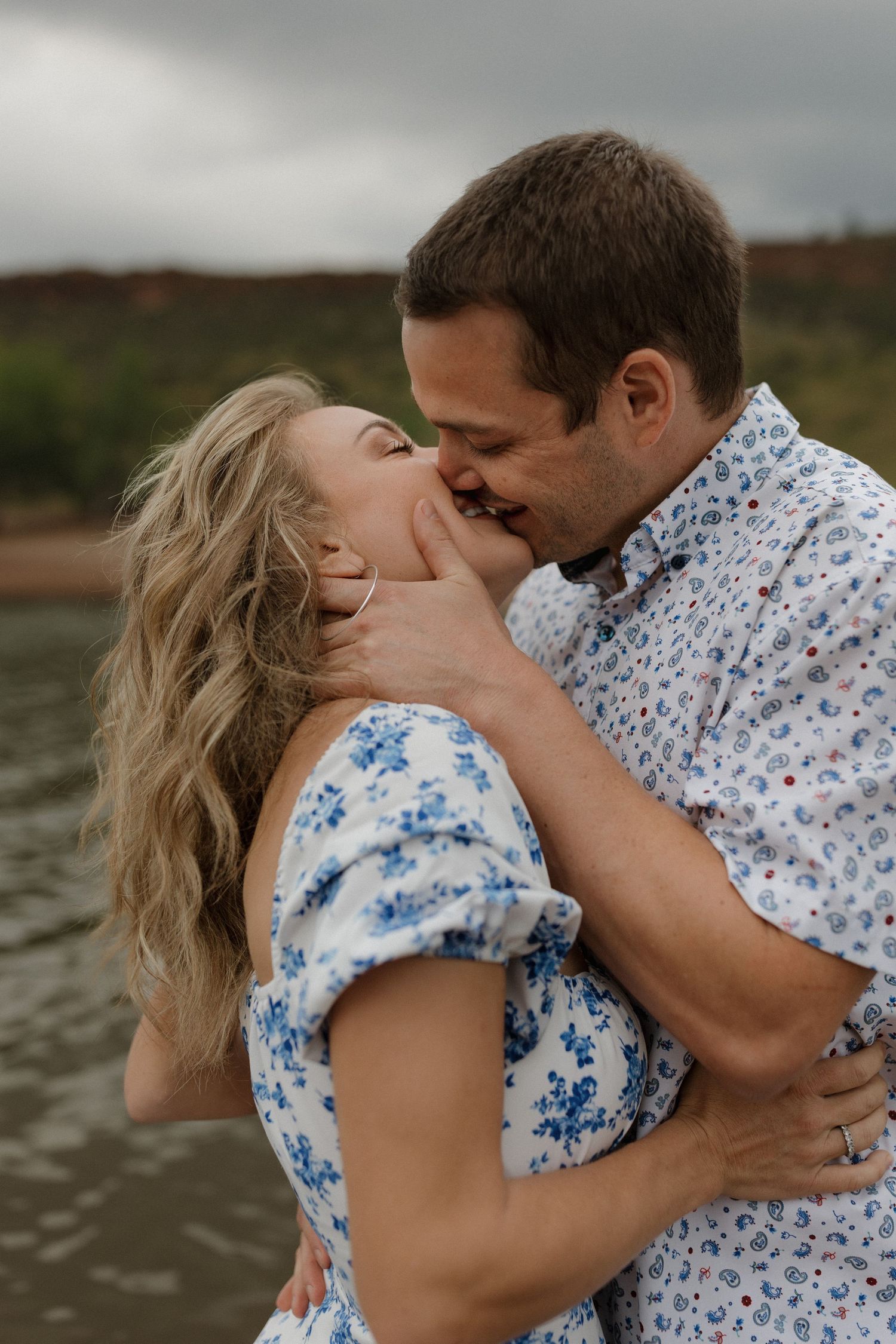 A couple shares a romantic kiss near a lake, dressed in matching blue floral outfits on a cloudy day.