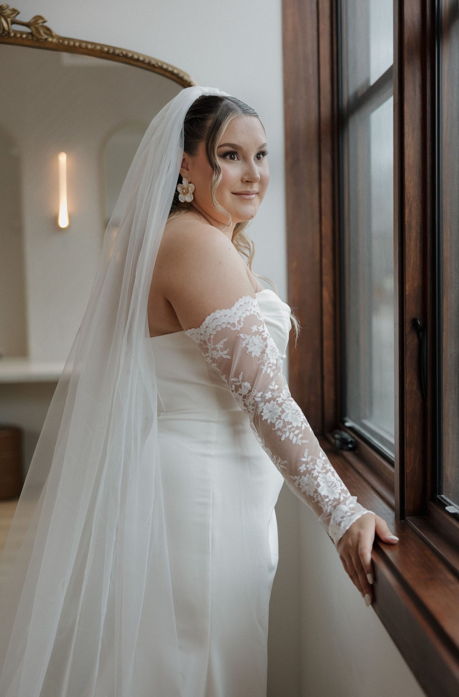 A bride in an elegant white lace-sleeved wedding dress and veil poses beside a window in natural light.