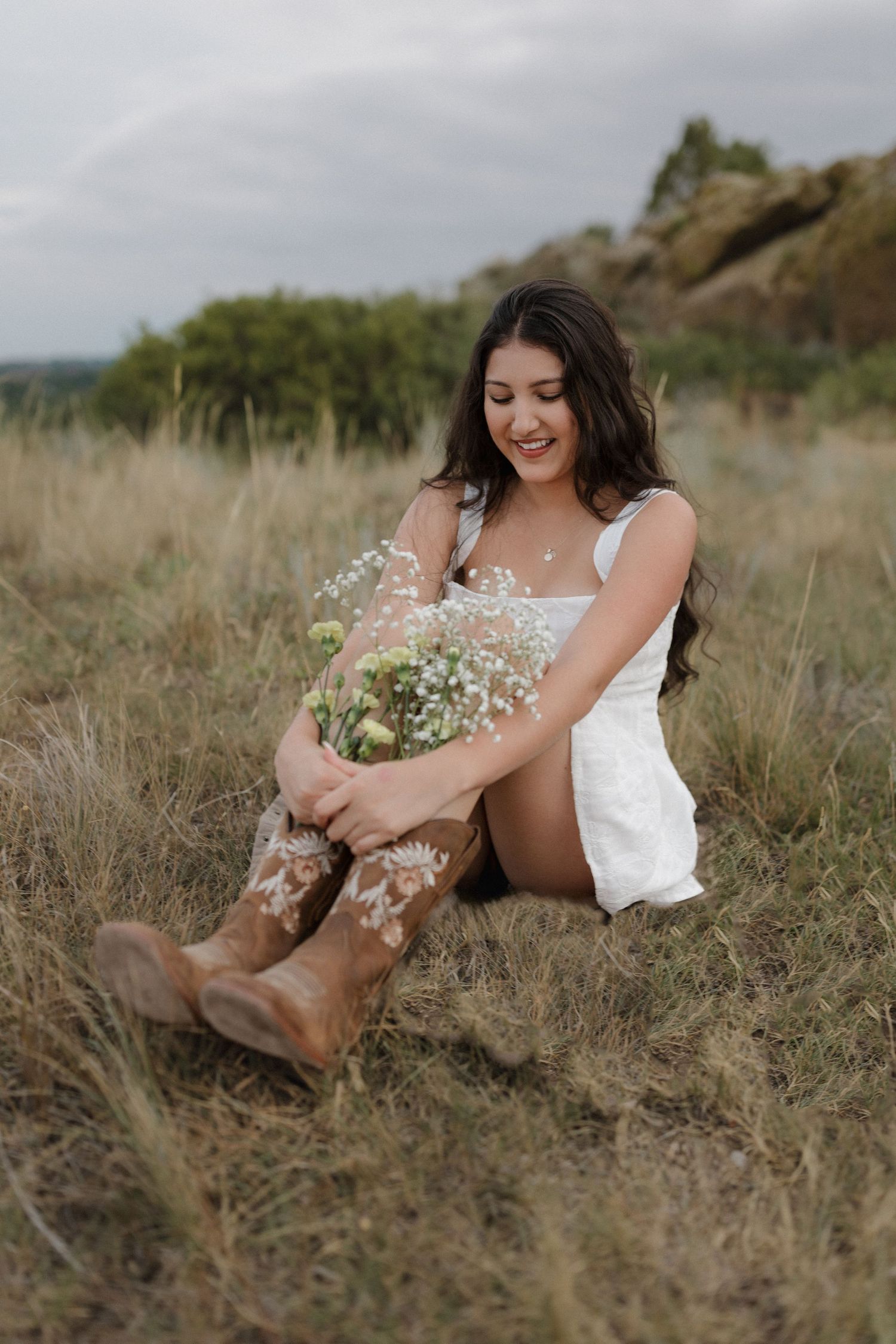 Person in a white dress sits barefoot in dried grass while holding delicate white wildflowers in a rustic outdoor setting.