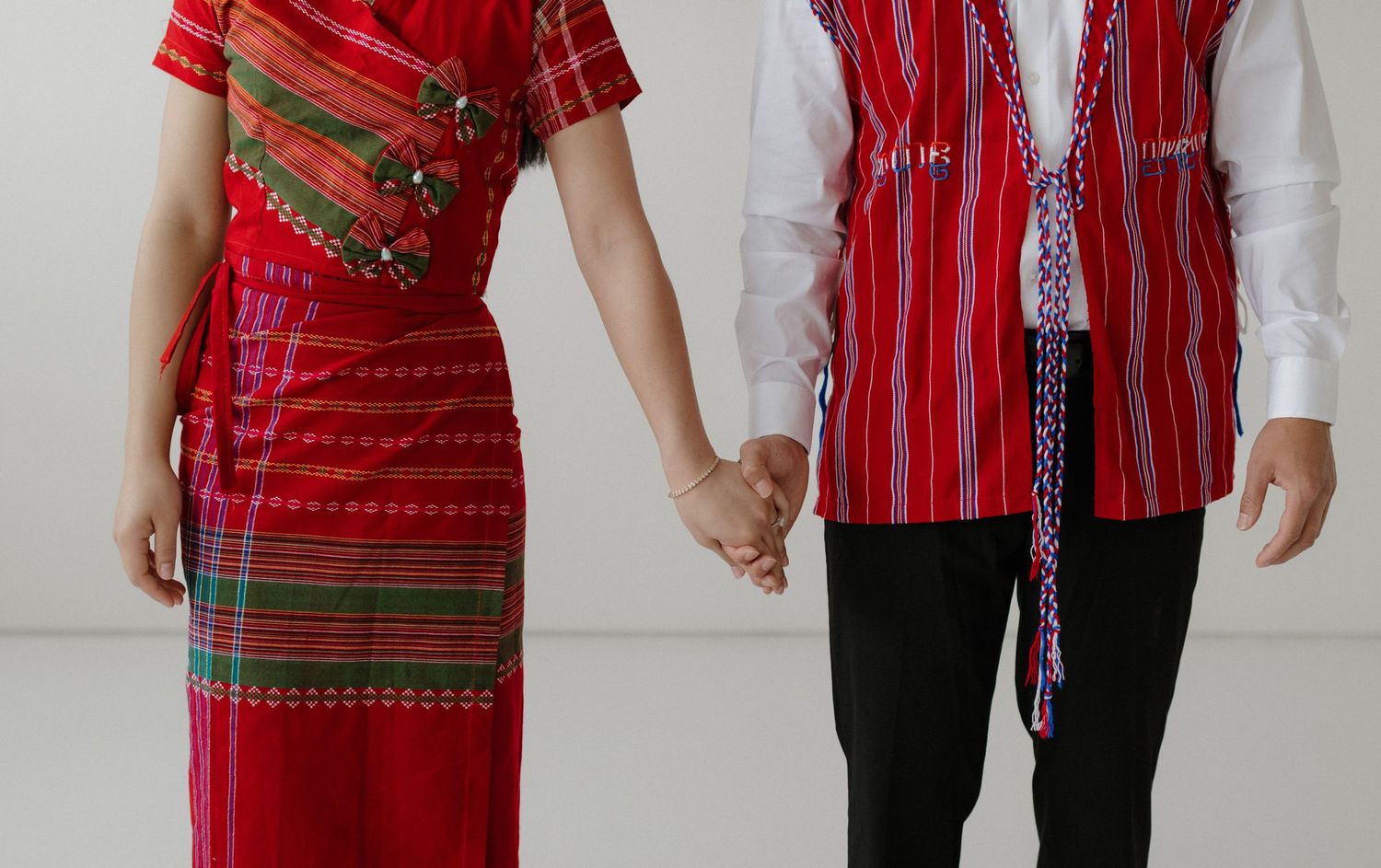 Couple wearing traditional red ethnic attire holding hands against white wall, showcasing cultural fashion.
