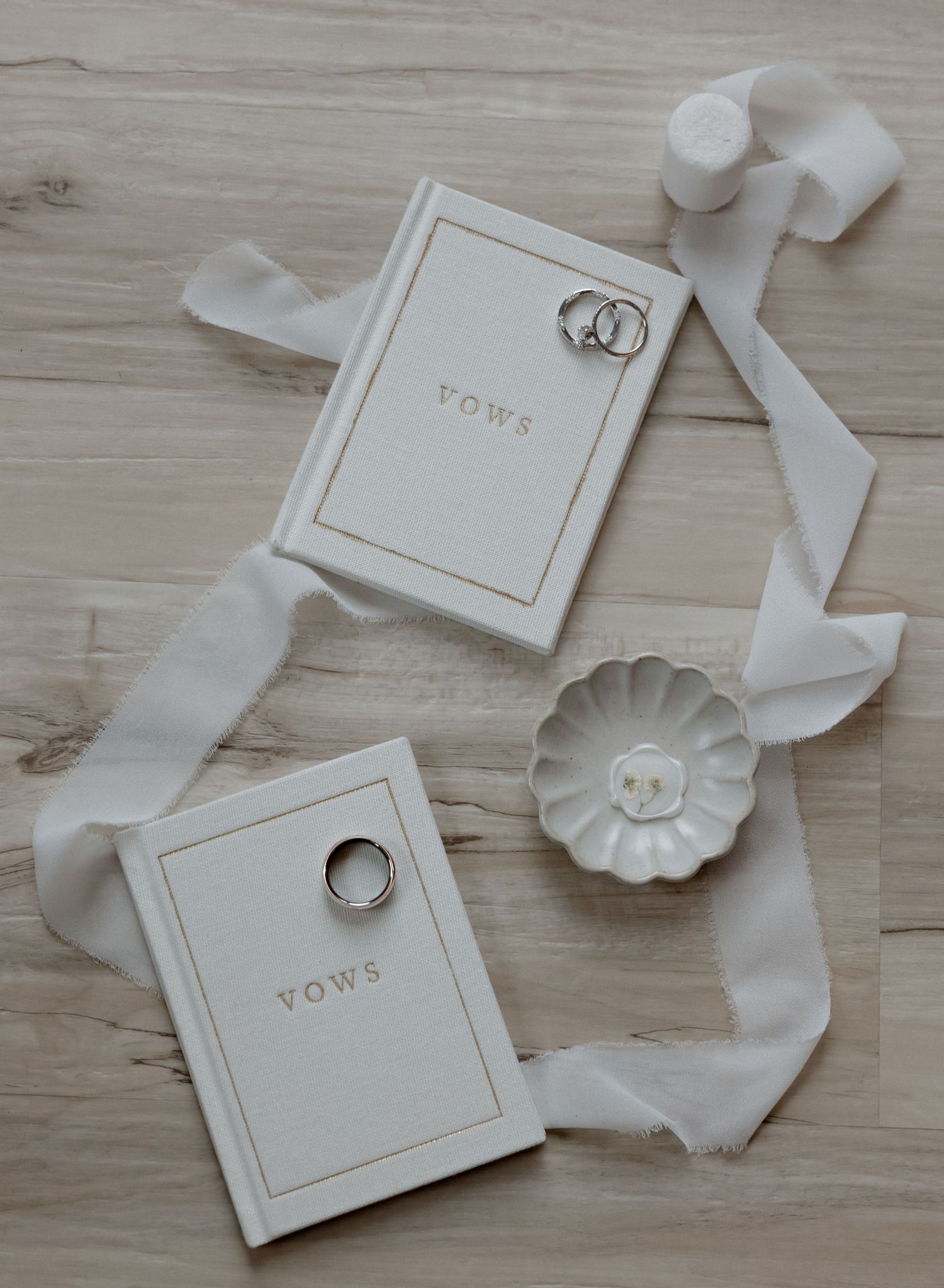 Two white vow books with wedding rings and delicate ribbon displayed on a light wooden surface with a small ceramic dish.