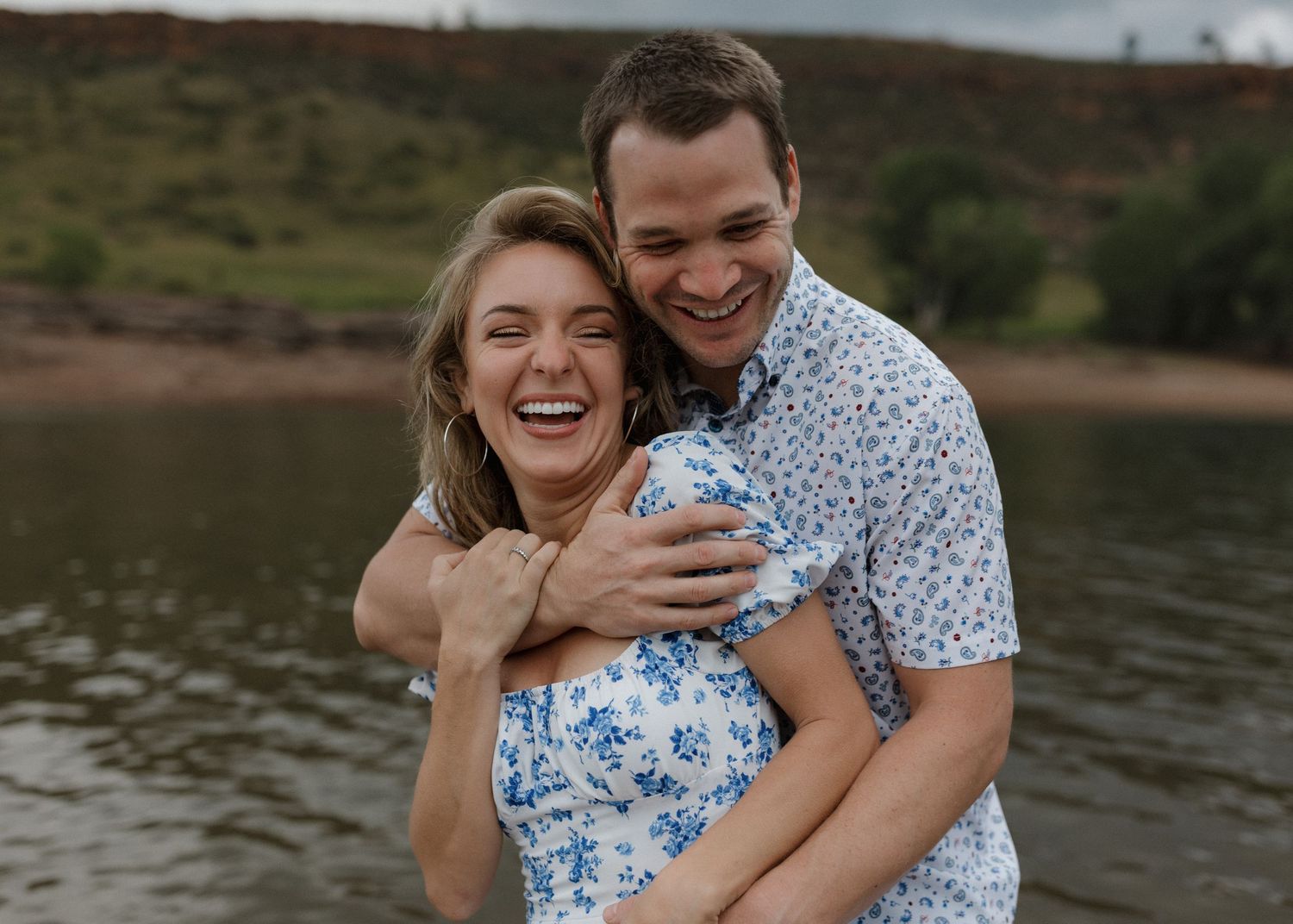 Affectionate couple in matching blue and white outfits laughing and embracing by a scenic lakeside.