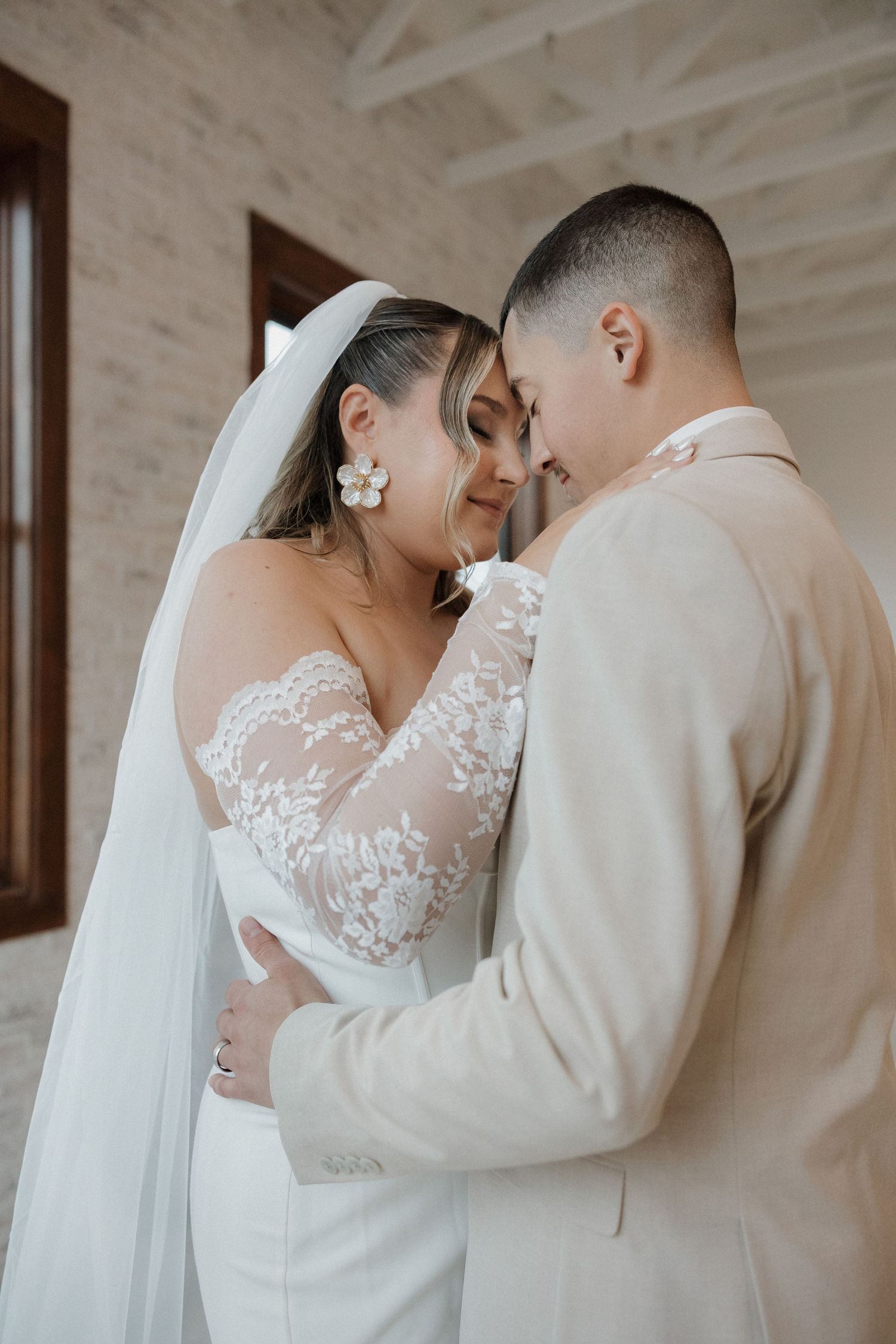A tender wedding moment between a bride in a lacy gown and groom in a beige suit sharing an intimate embrace.