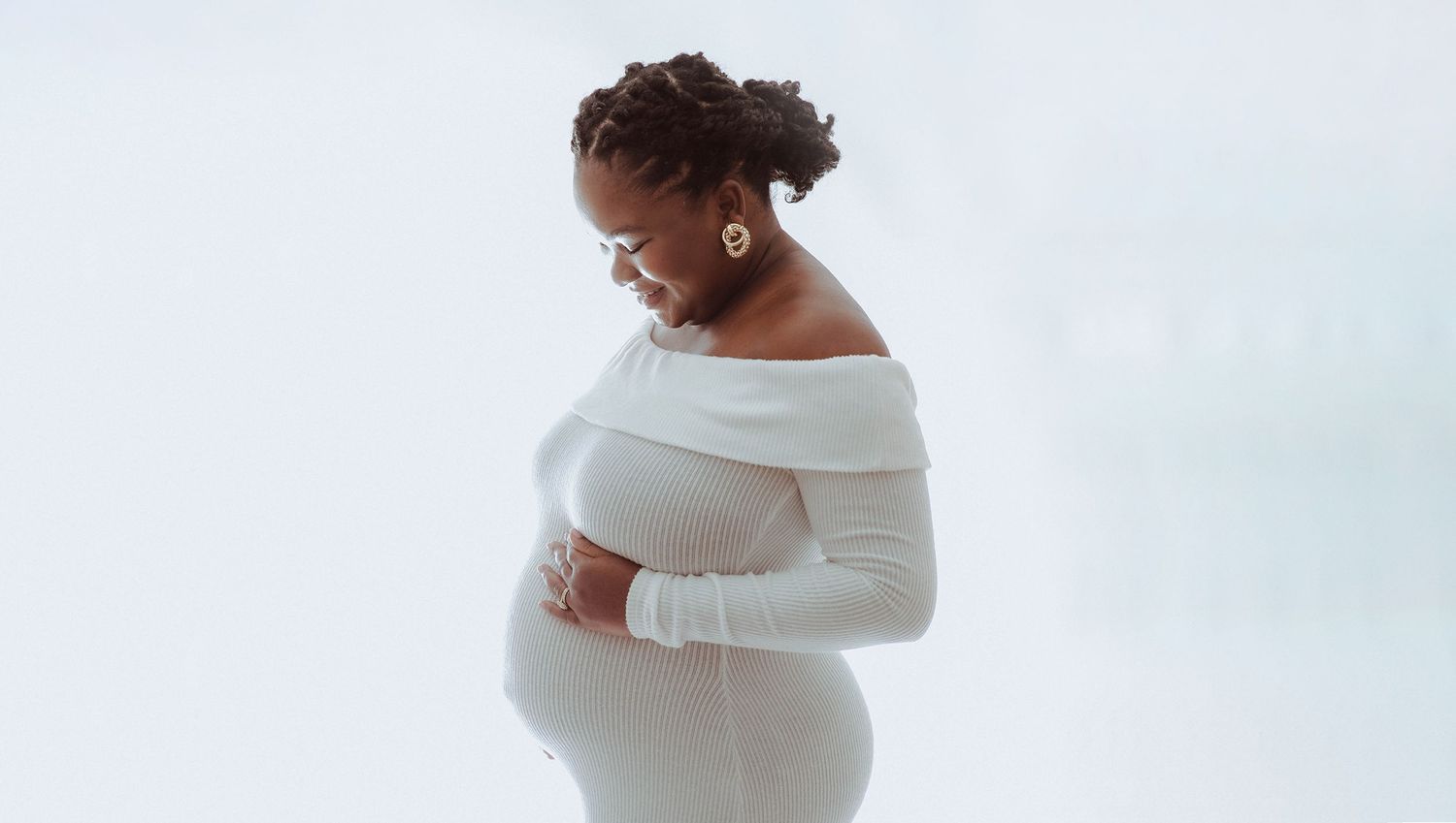 Elegant maternity photo of an expecting mother wearing a white dress against a light background. During a maternity session in Bexley