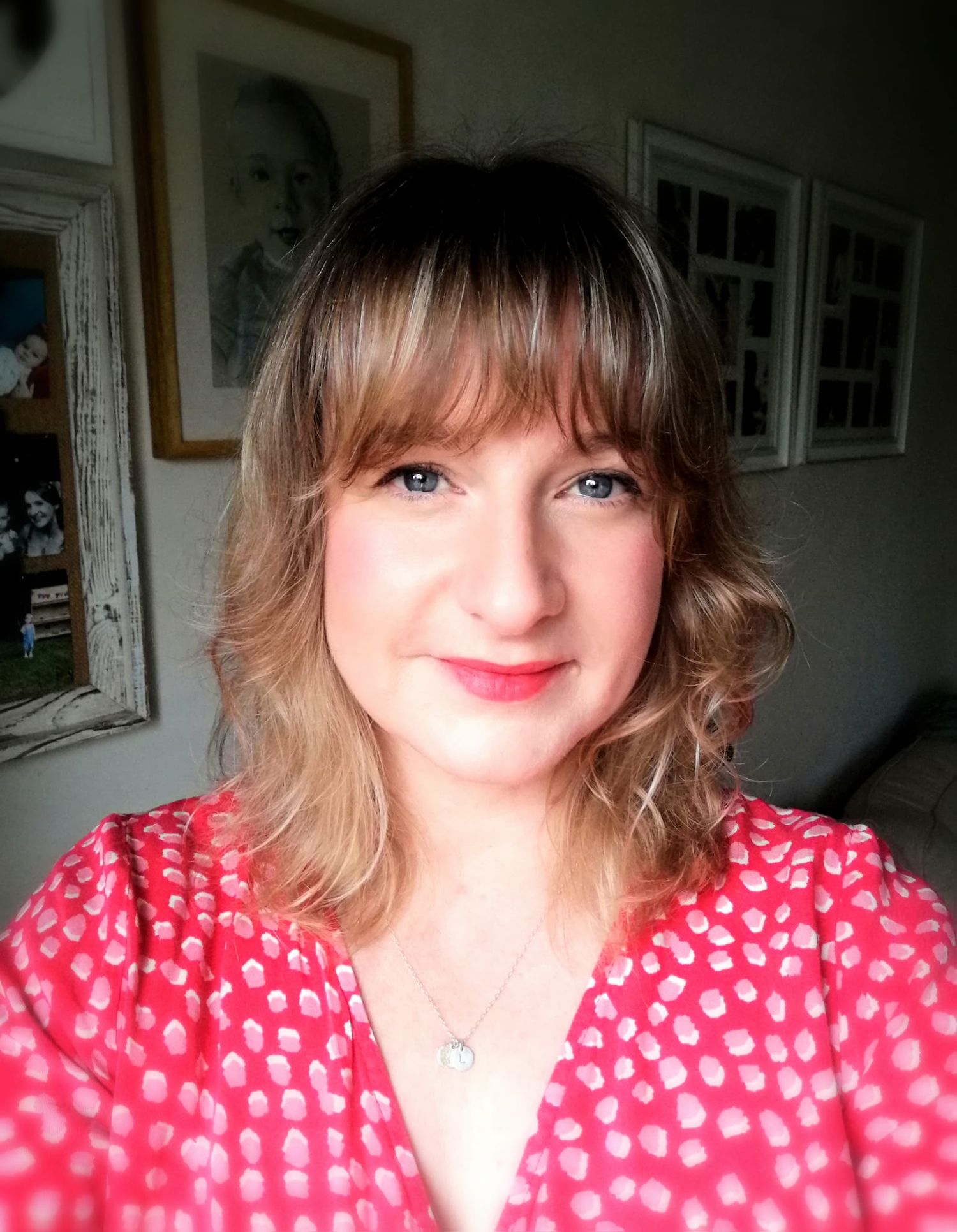 Smiling woman with blonde bangs wearing a red polka dot top and silver necklace, posing indoors with framed art.