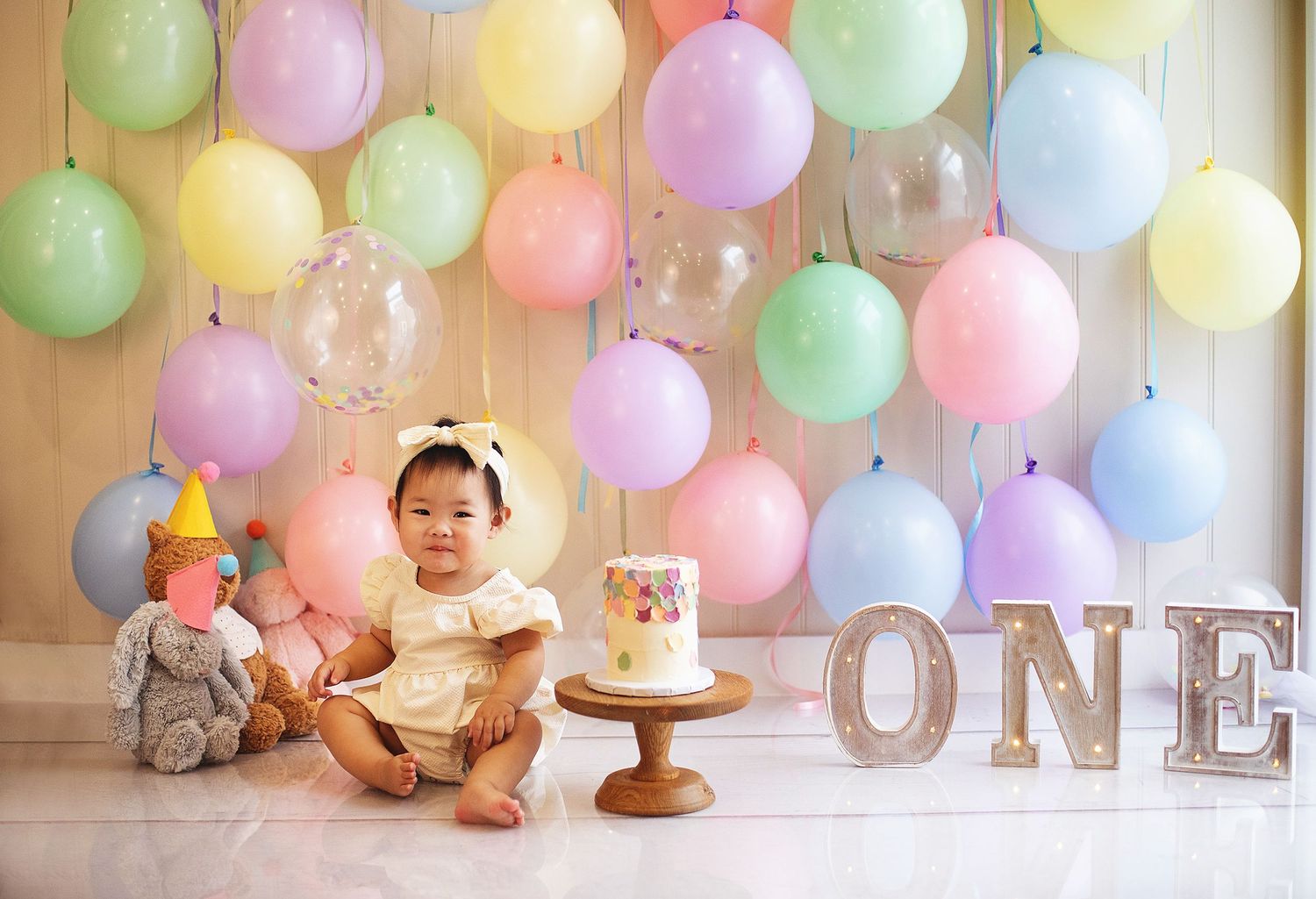 Baby girl in yellow dress celebrates first birthday with colorful balloons, cake, and ONE letters.