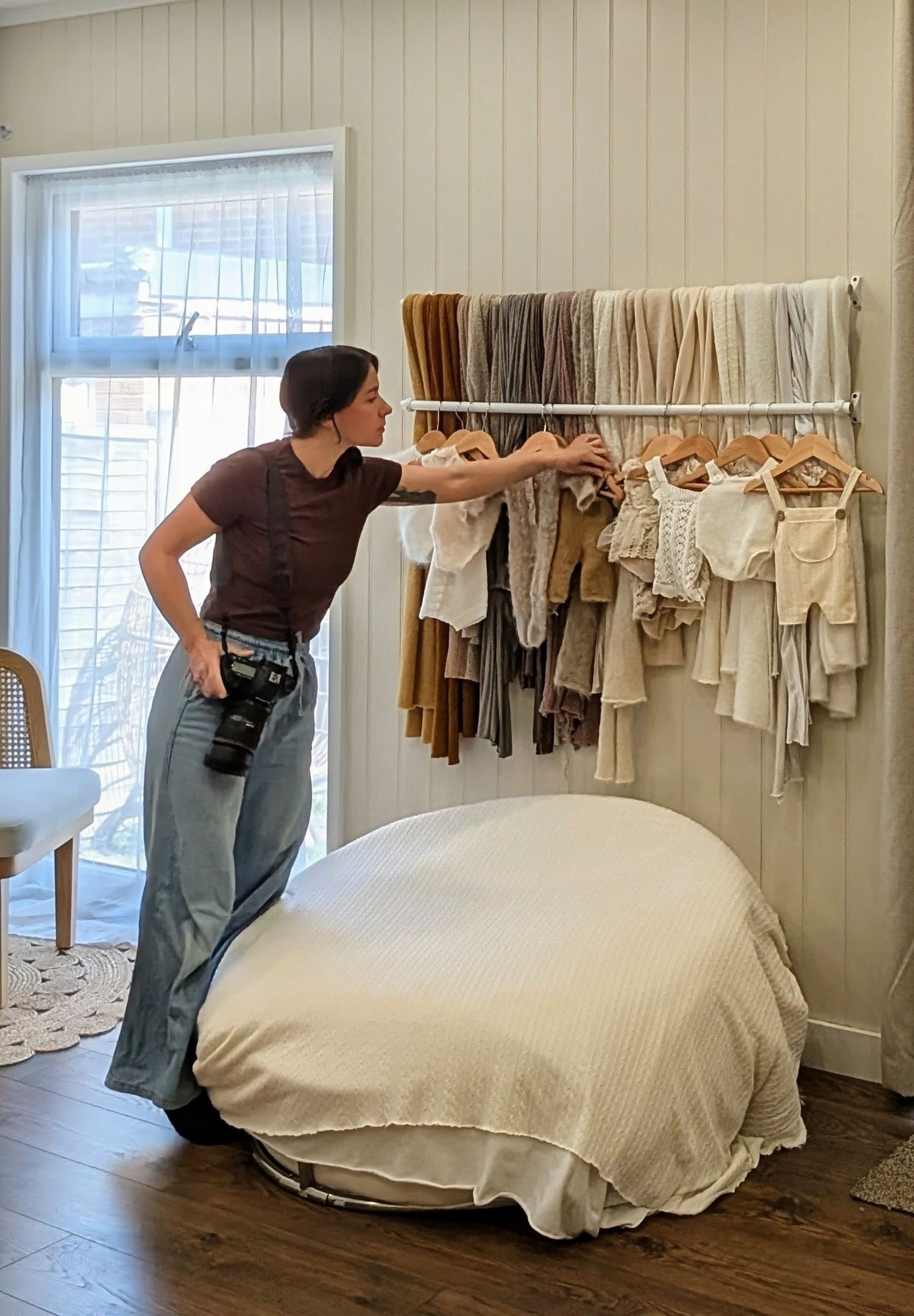 newborn Photographer browses neutral-toned baby clothing on a rack beside a draped posing bed in a bright photography studio in Bexley