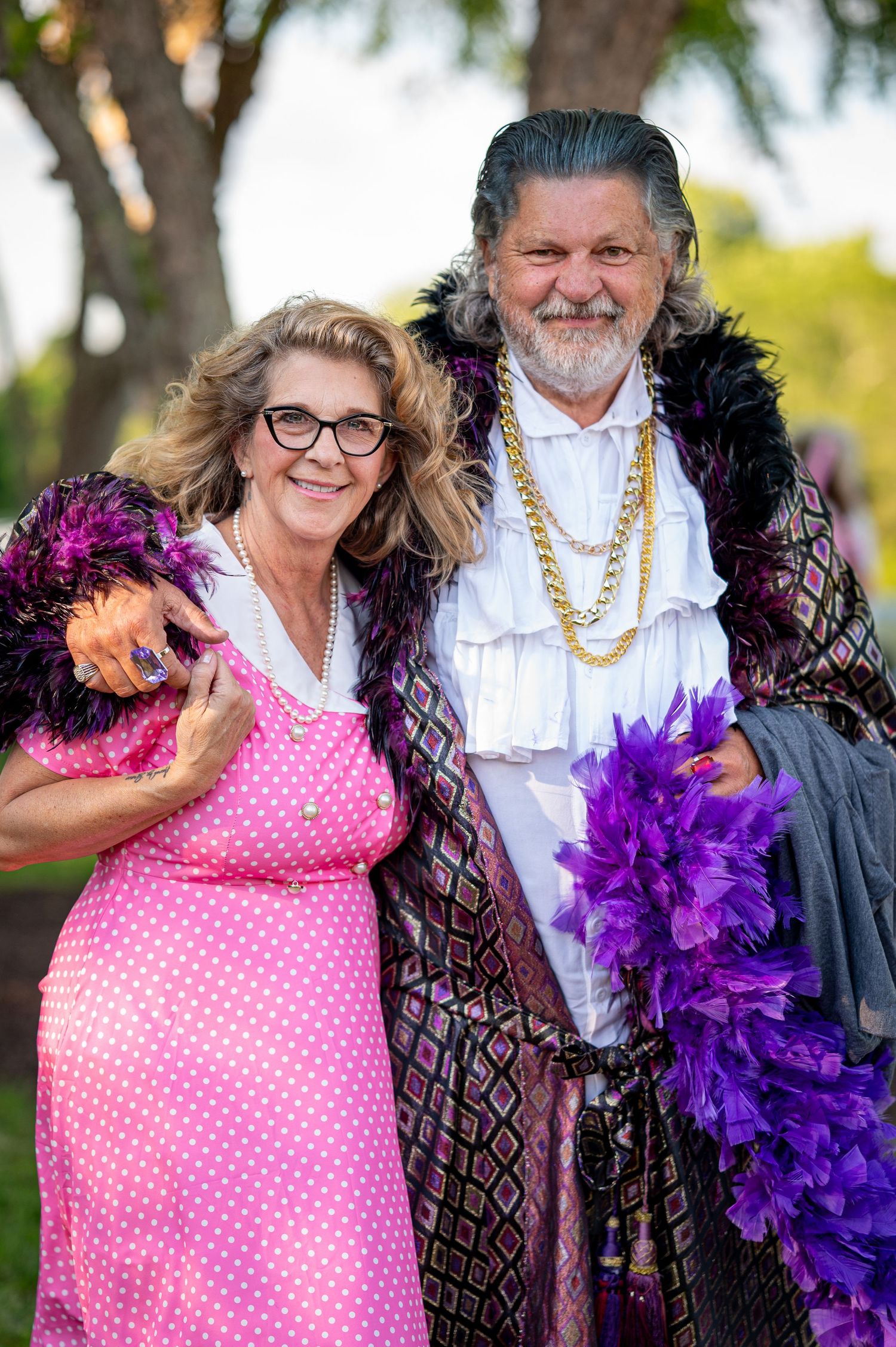 A festively dressed couple in polka dots and royal attire holding purple and pink feather boas.