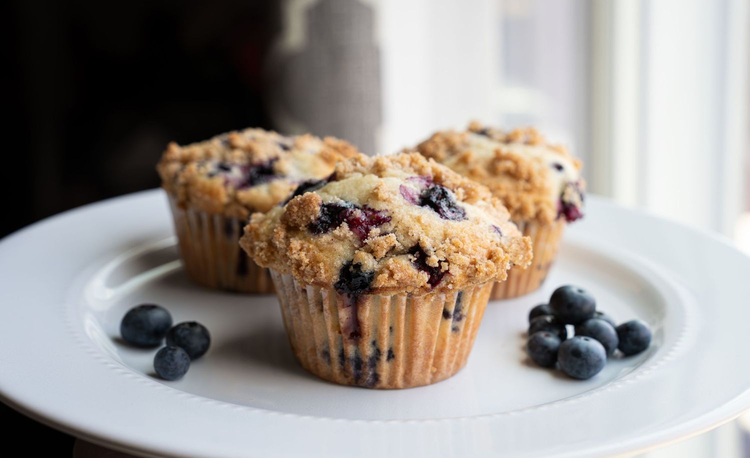 Fresh baked blueberry muffins with streusel topping on white plates with scattered fresh blueberries.