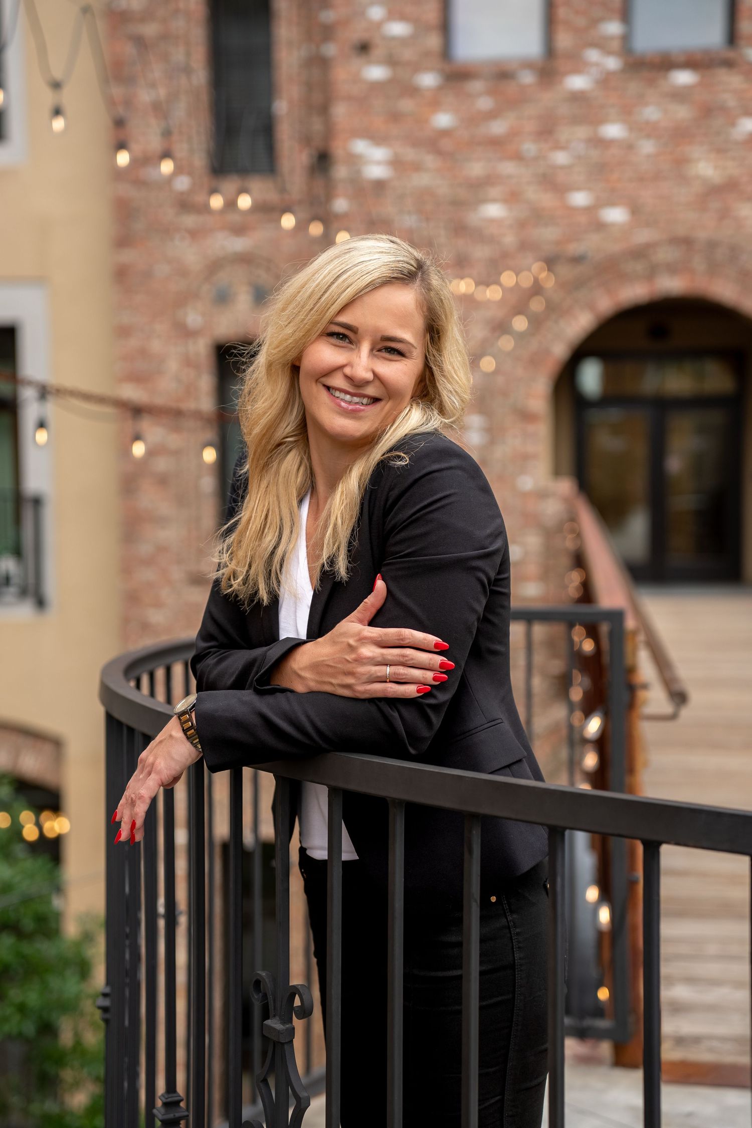 Person in black business attire leans on outdoor railing with exposed brick wall and string lights in background.