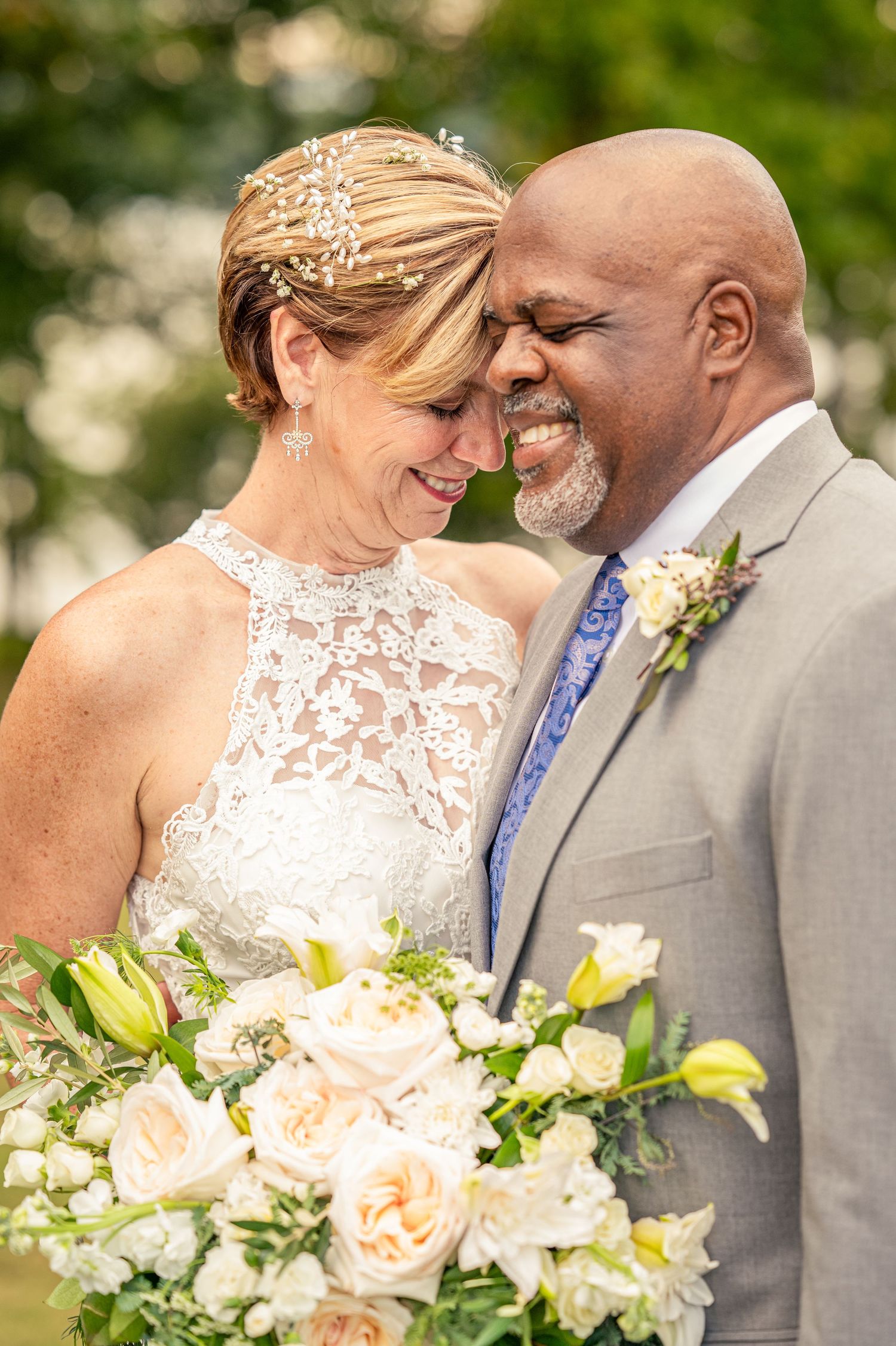 A joyful couple in wedding attire share an intimate moment while holding a bouquet of peach roses outdoors.