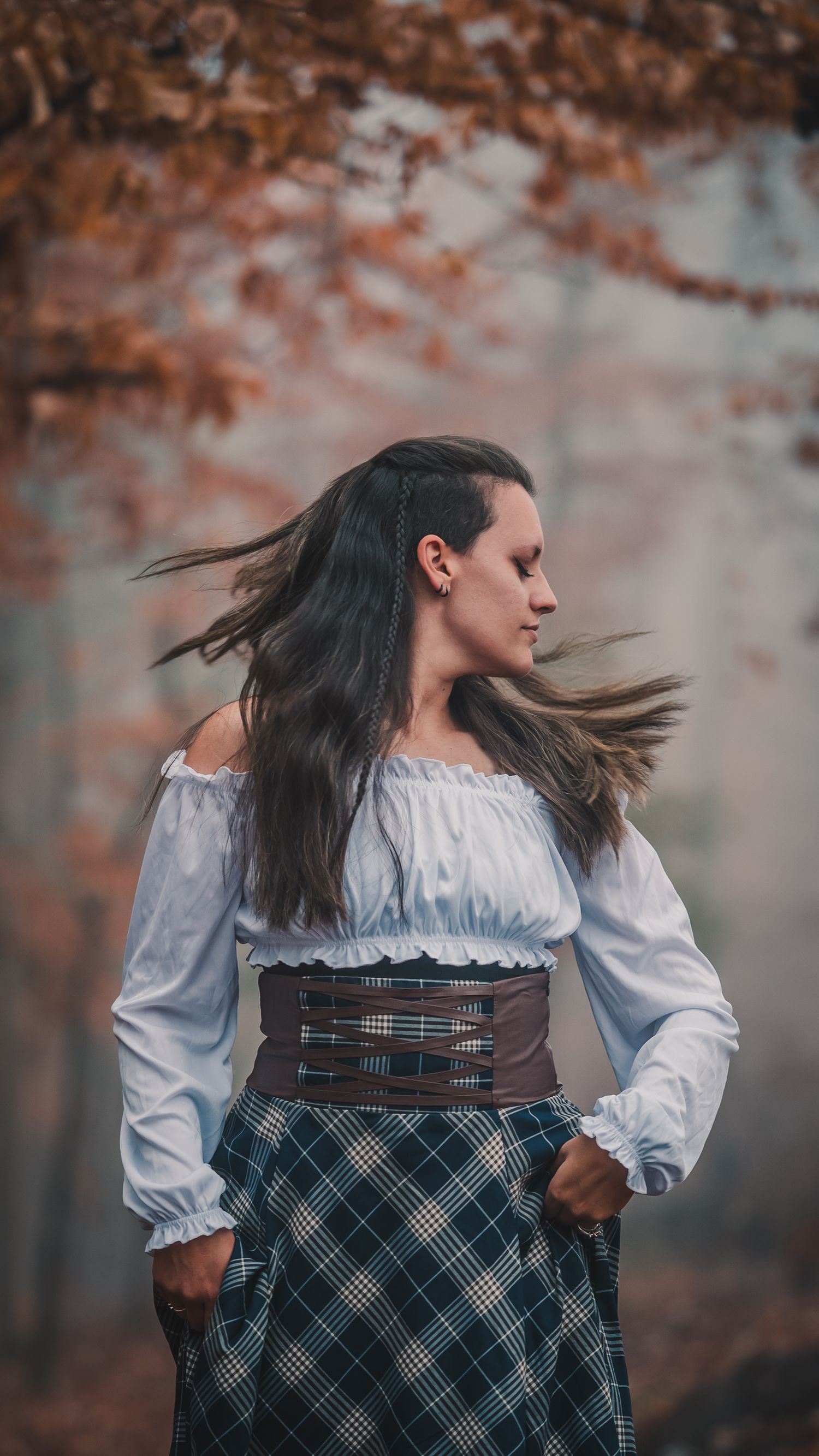 A woman in a historical costume with plaid skirt and corset twirls against an autumn forest background.