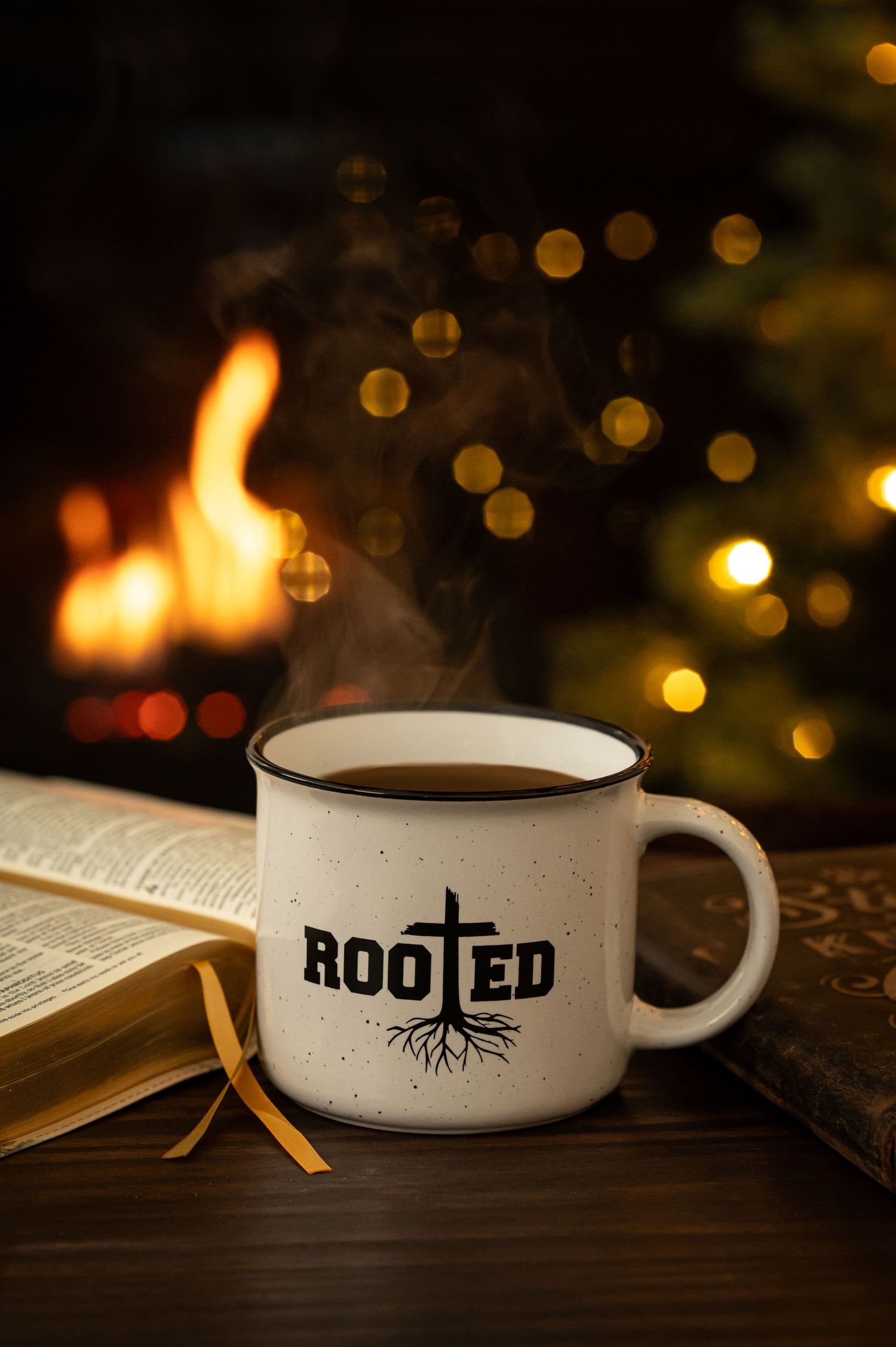 A steaming Rooted mug sits beside an open book with a cozy fireplace in the background.
