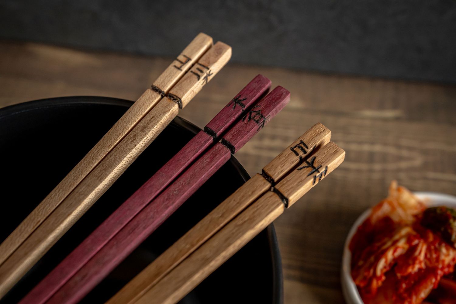 Three pairs of wooden chopsticks in natural and burgundy colors rest beside a dark bowl on a rustic wooden surface.