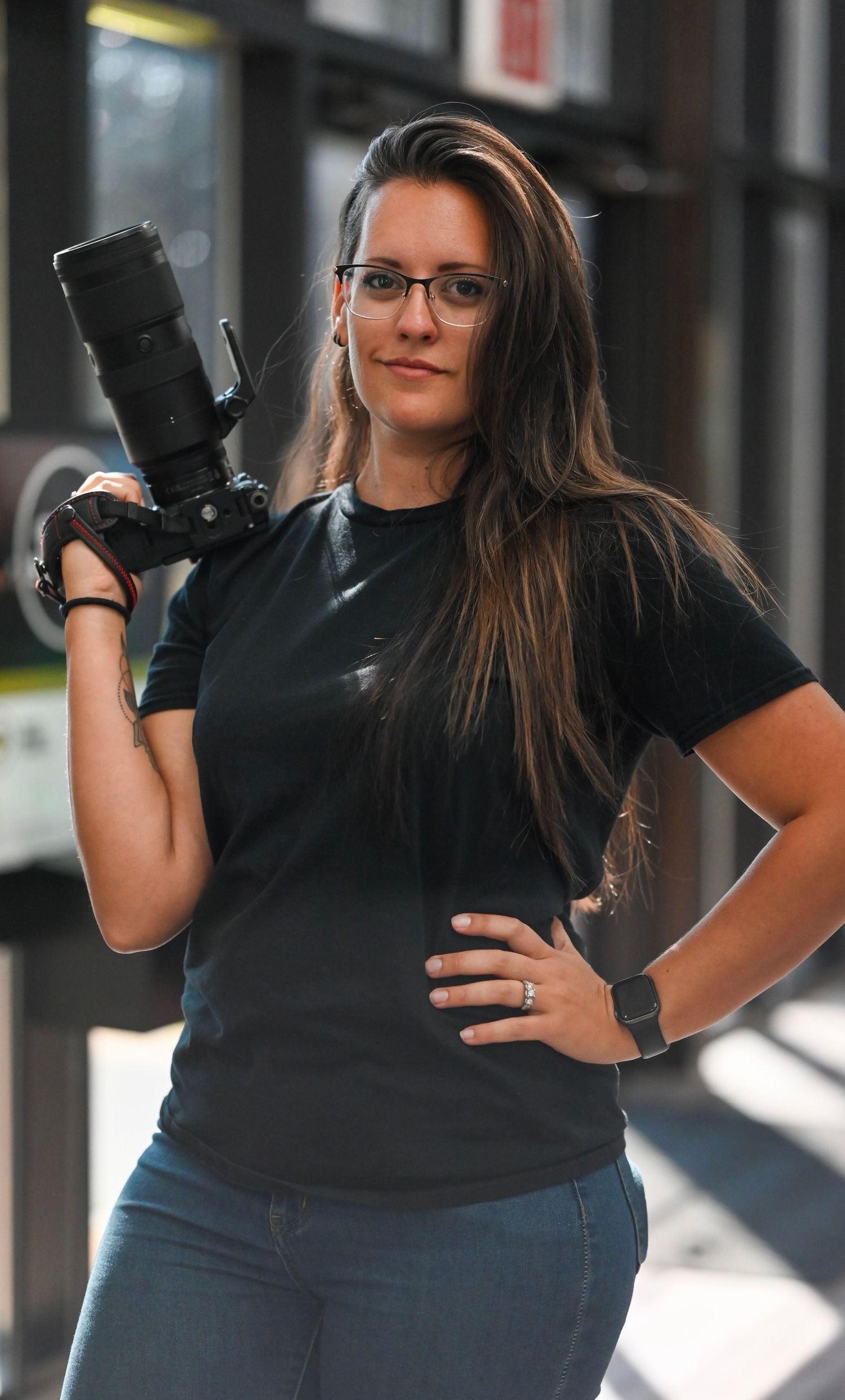 Professional photographer in a black t-shirt holds a DSLR camera with telephoto lens against an urban background.