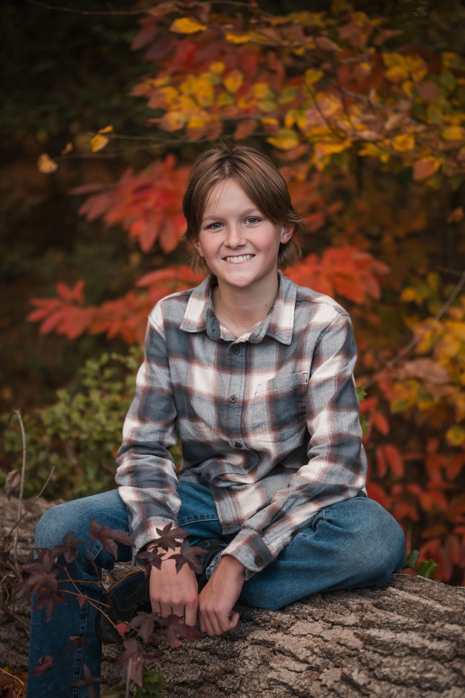 Autumn portrait session featuring a plaid flannel shirt and jeans against vibrant fall foliage and a rustic log.