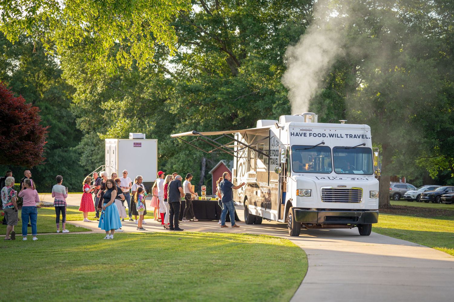A food truck festival in a sunny park with vendors and customers gathering on a grassy lawn.