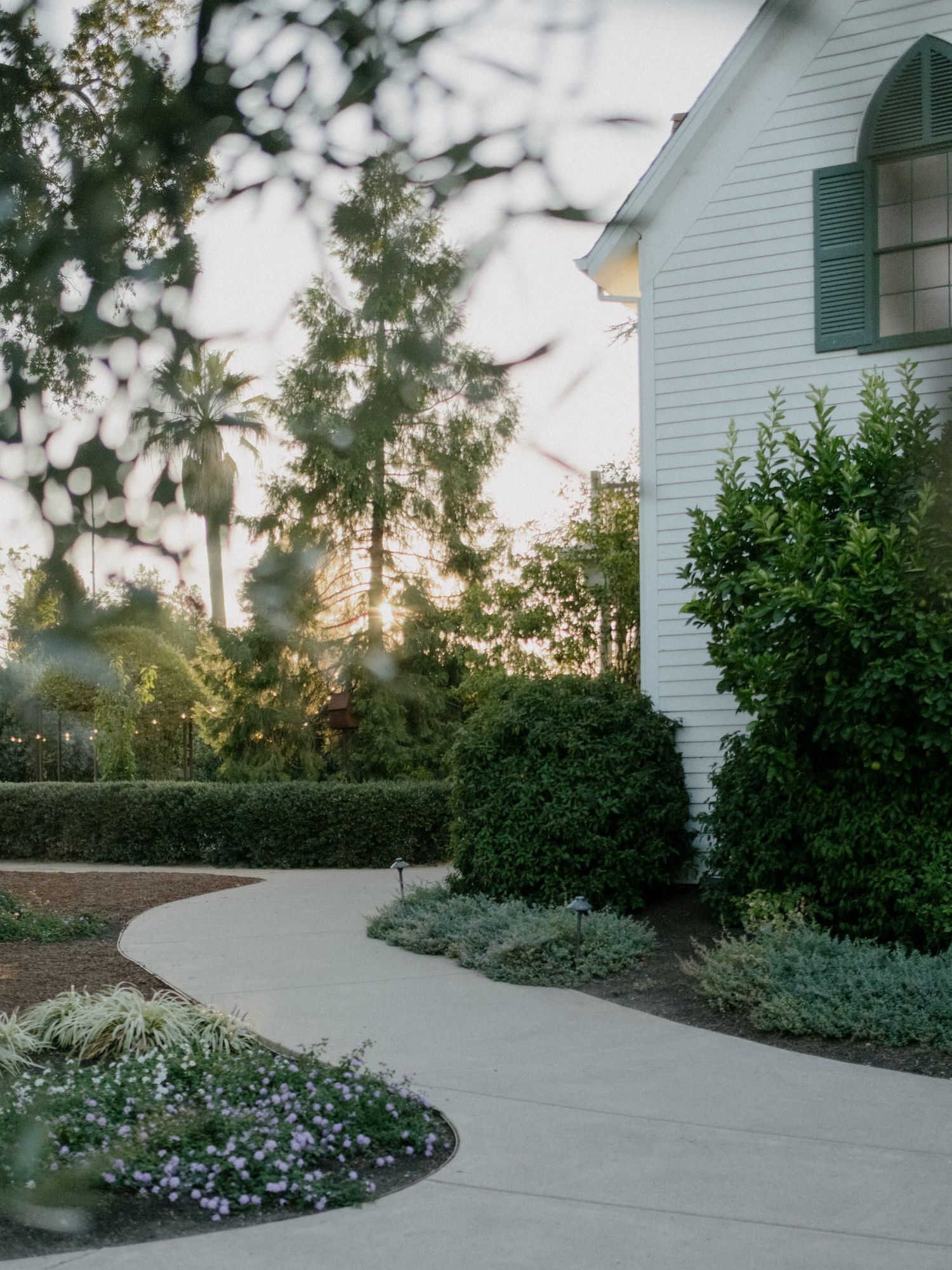 White chapel with green shutters surrounded by lush hedges and flowering plants along a curved concrete pathway at sunset.