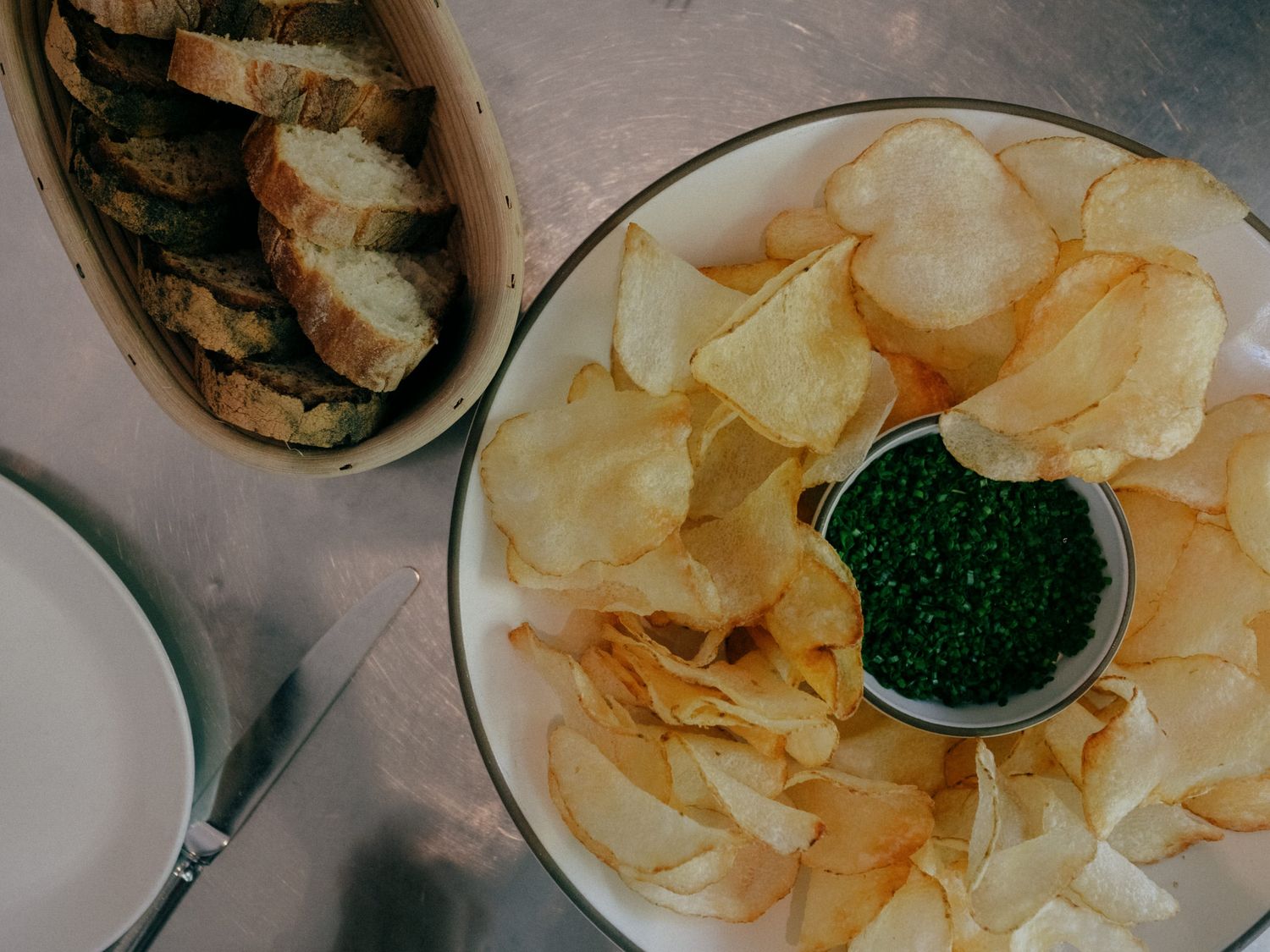A plate of crispy potato chips surrounding a small bowl of green caviar, with sliced bread in a basket nearby.