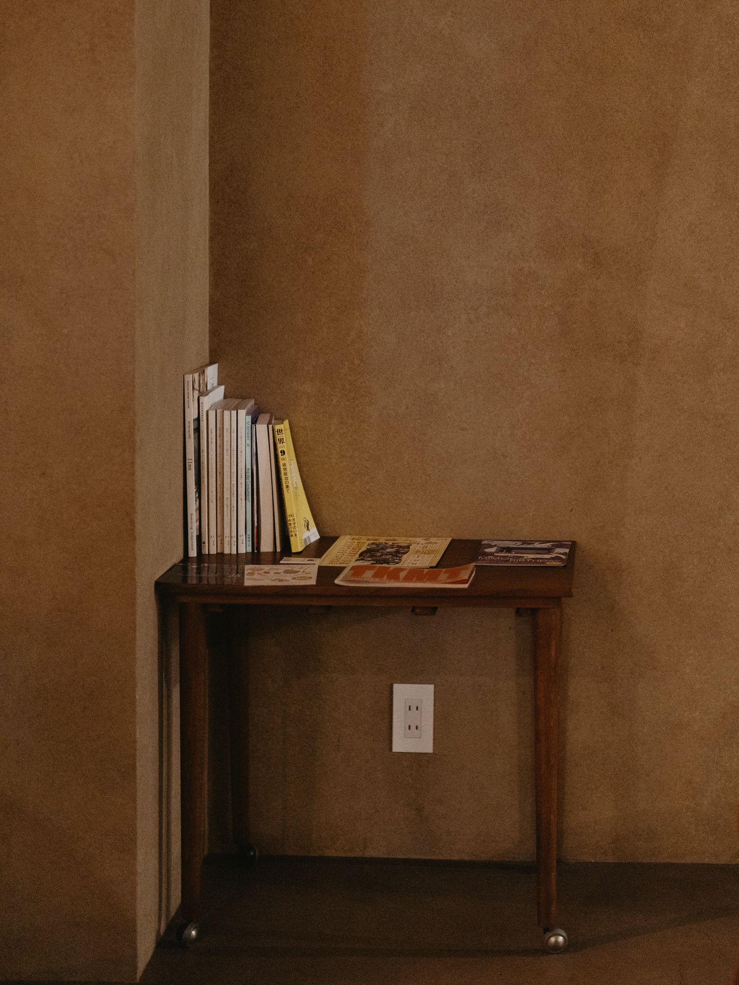 Wooden shelf mounted on brown wall with electrical outlet and scattered books creating minimalist decor.