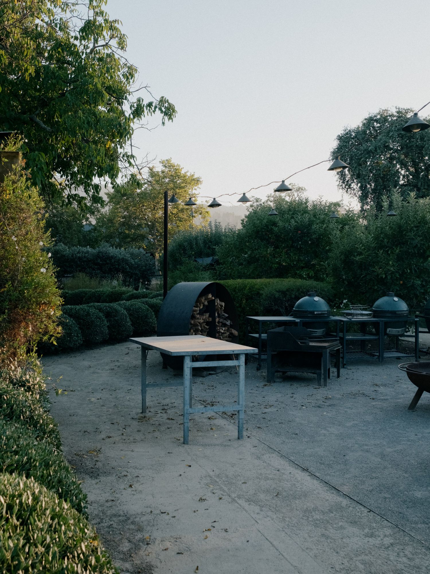 Outdoor garden patio with a white table, green egg grills, string lights, and lush hedges at dusk.
