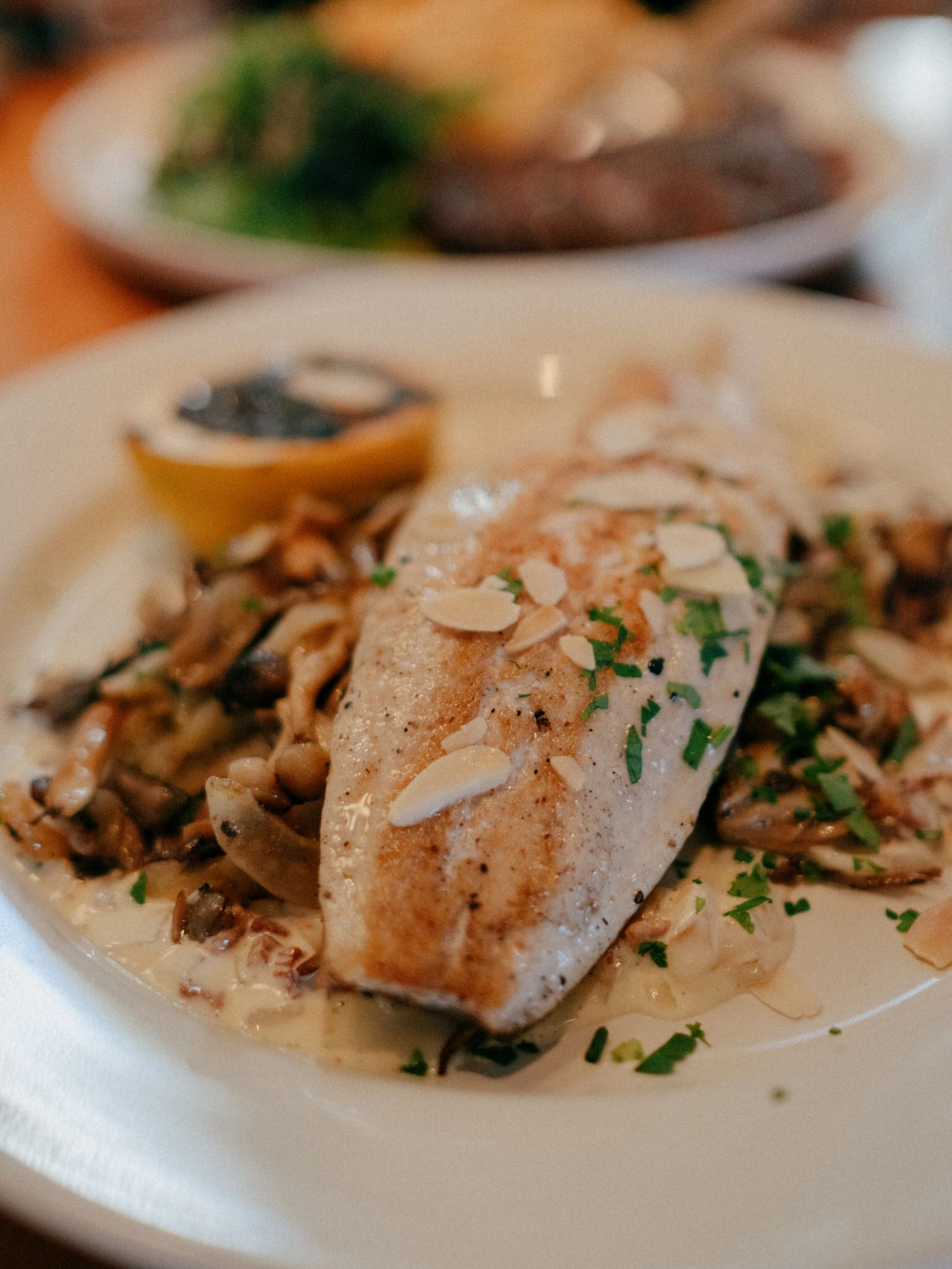 Grilled fish fillet topped with toasted almonds and herbs, served with sautéed mushrooms and a lemon half on a white plate.