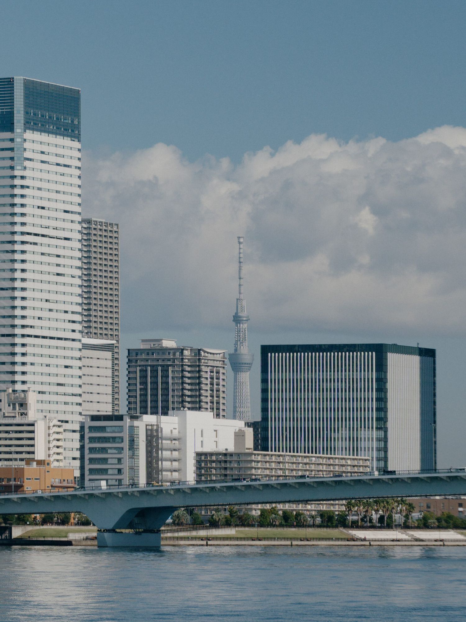 Modern waterfront cityscape with white high-rise buildings and bridge spanning across calm blue waters under cloudy sky.