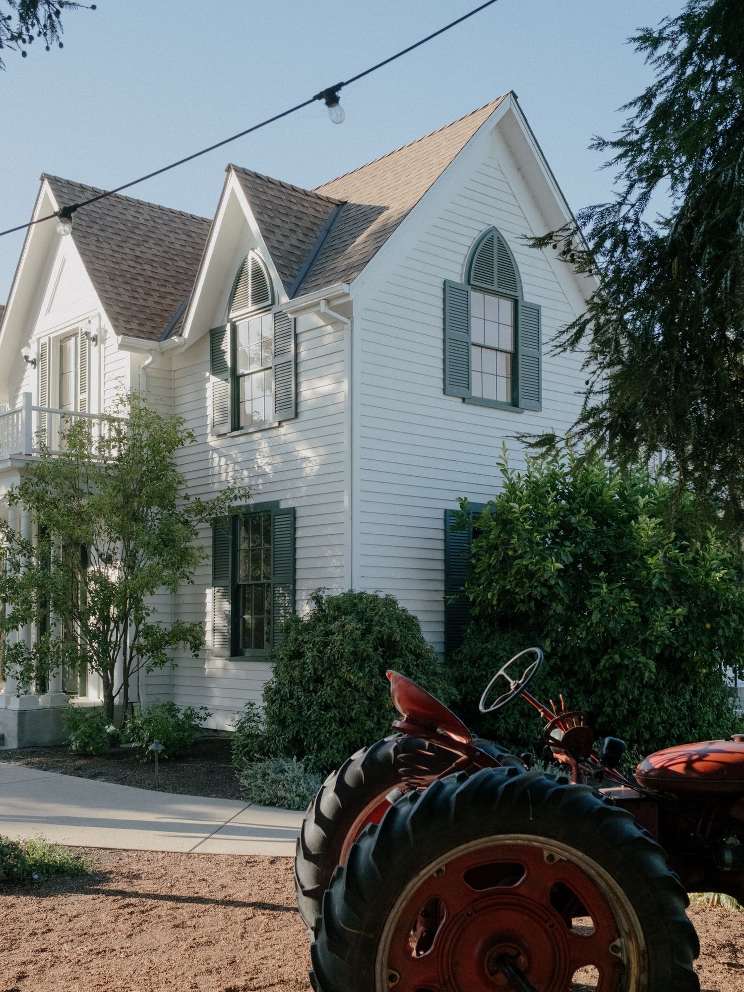 Red tractor parked in front of a charming white two-story farmhouse with dark shutters and lush green trees.
