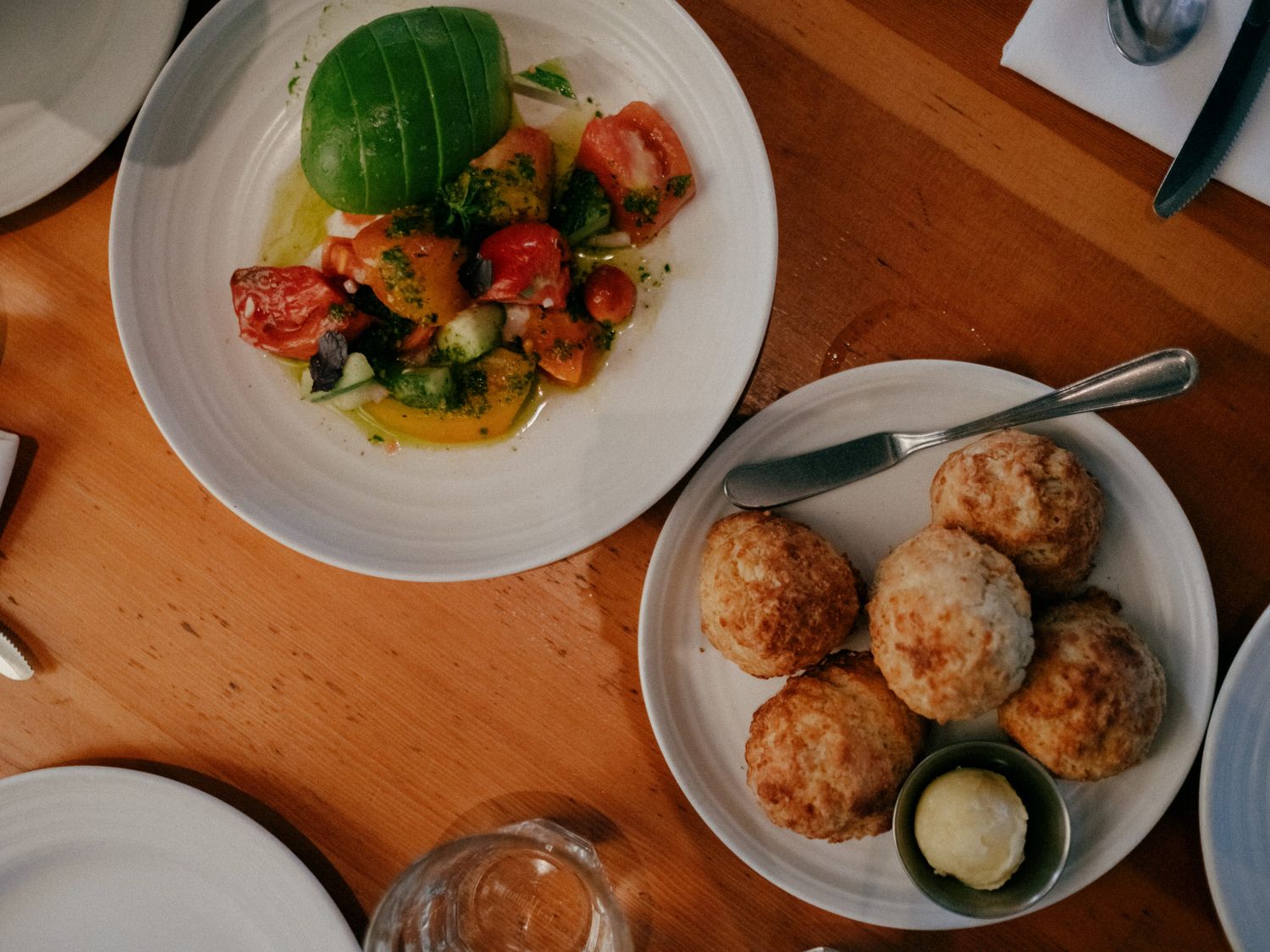 Overhead view of two white plates on wooden table, one with tomato salad and one with golden baked rolls and butter.