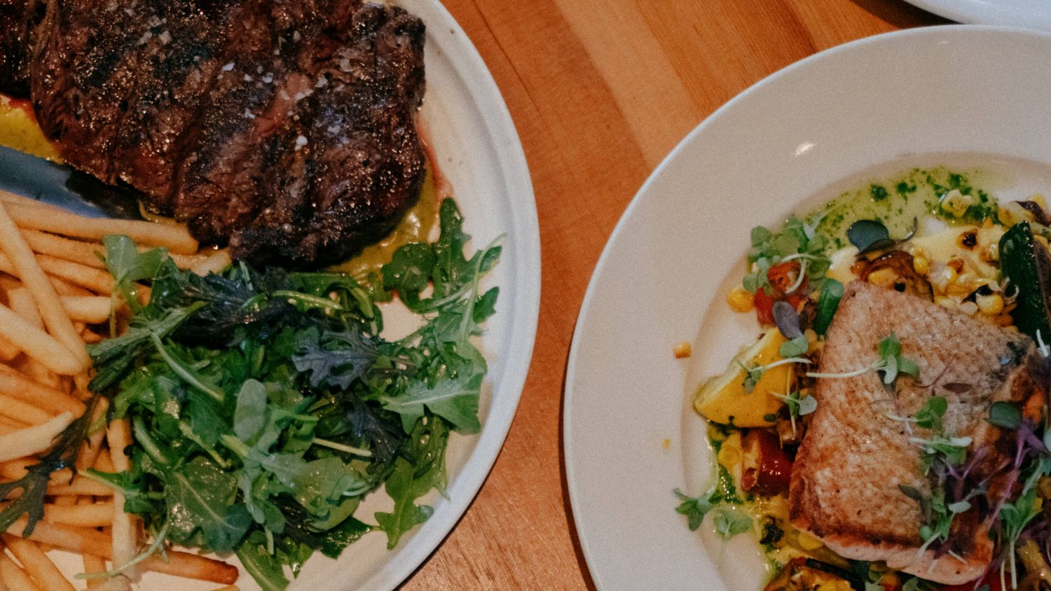 Two restaurant plates featuring grilled steak with fries and arugula salad beside a fresh salmon dish with colorful vegetables.