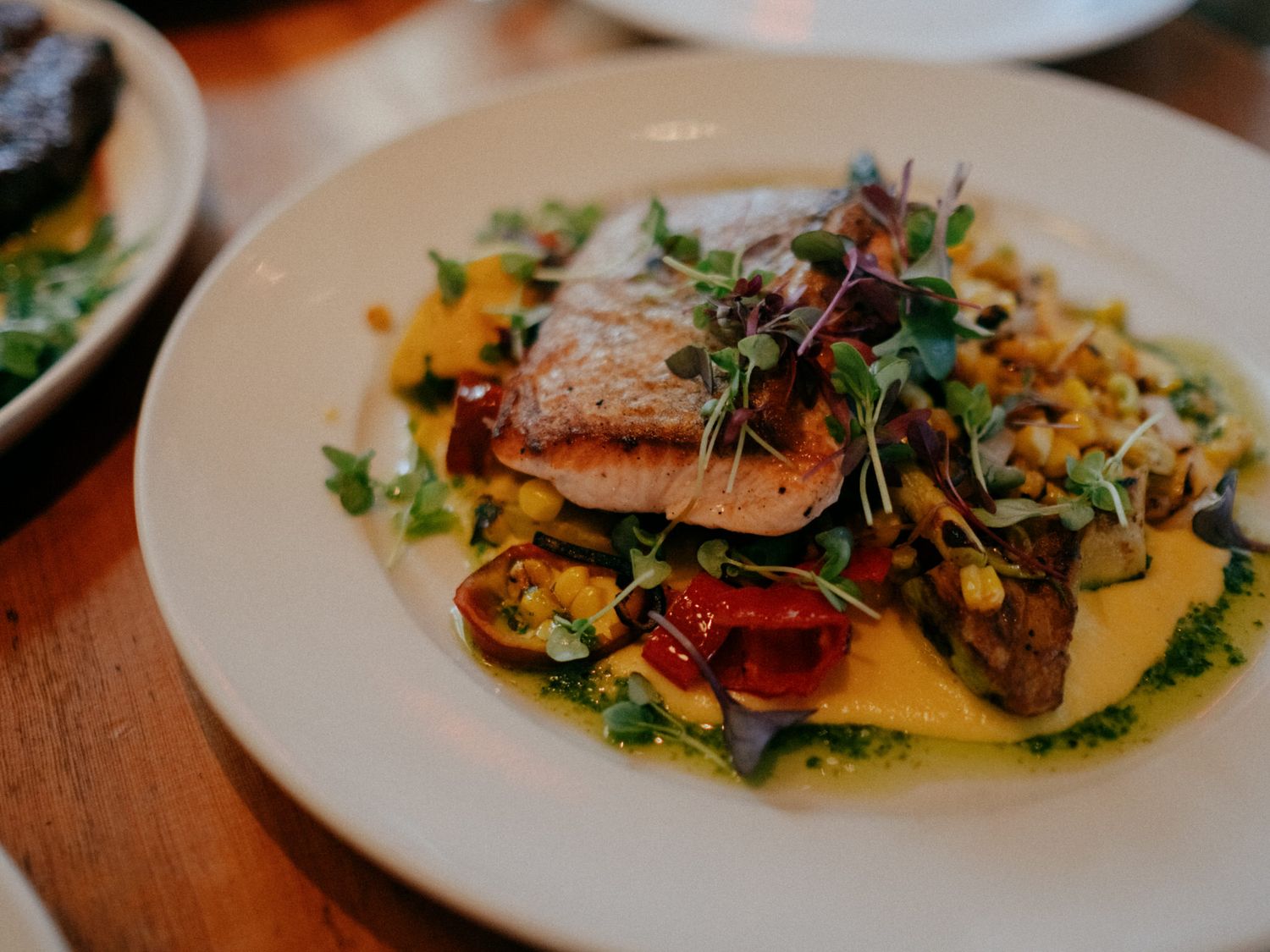 Seared salmon fillet served over roasted vegetables and creamy polenta, garnished with fresh microgreens and herb sauce.
